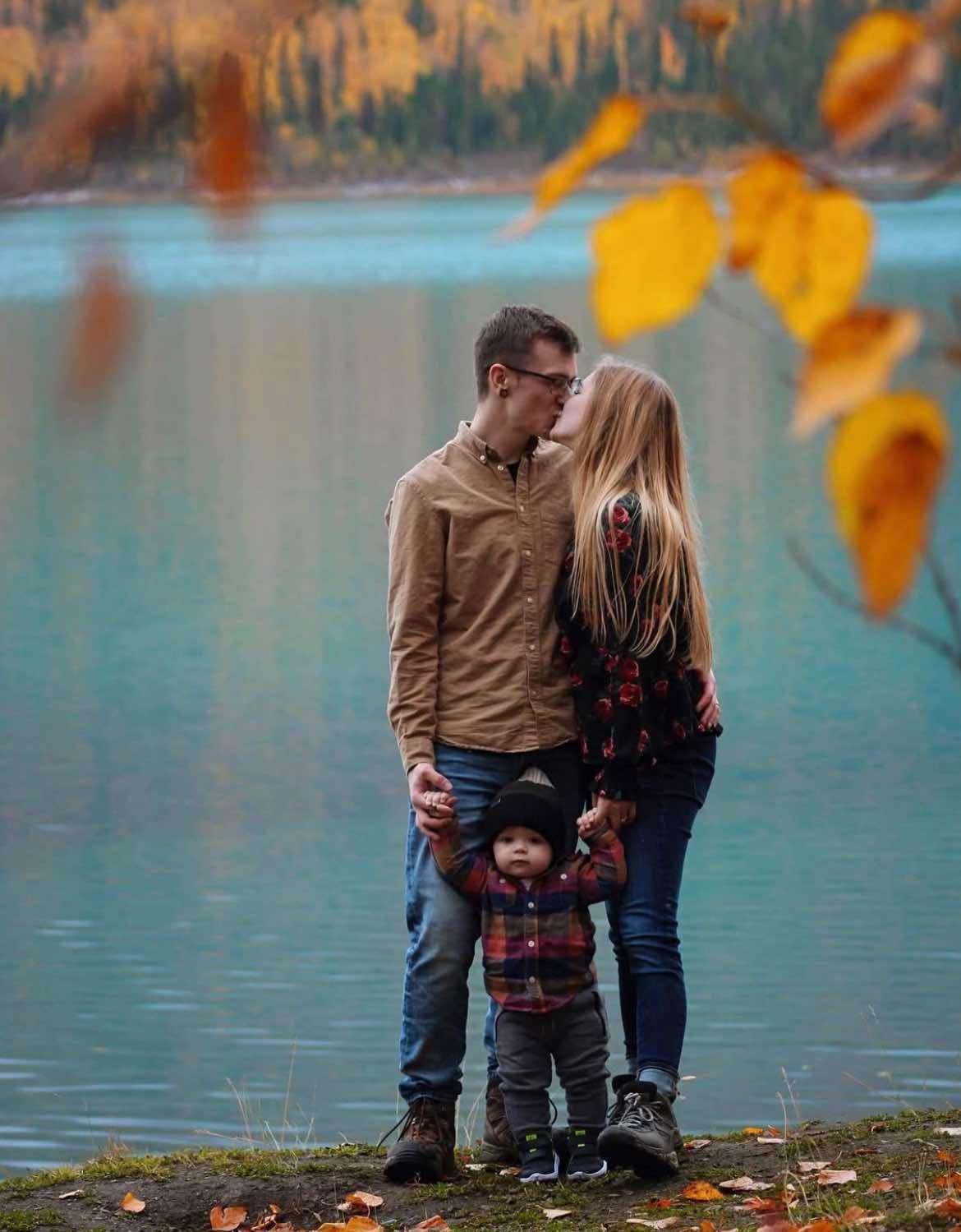 A couple and small child stand by a turquoise lake with autumn leaves framing the scene during fall.