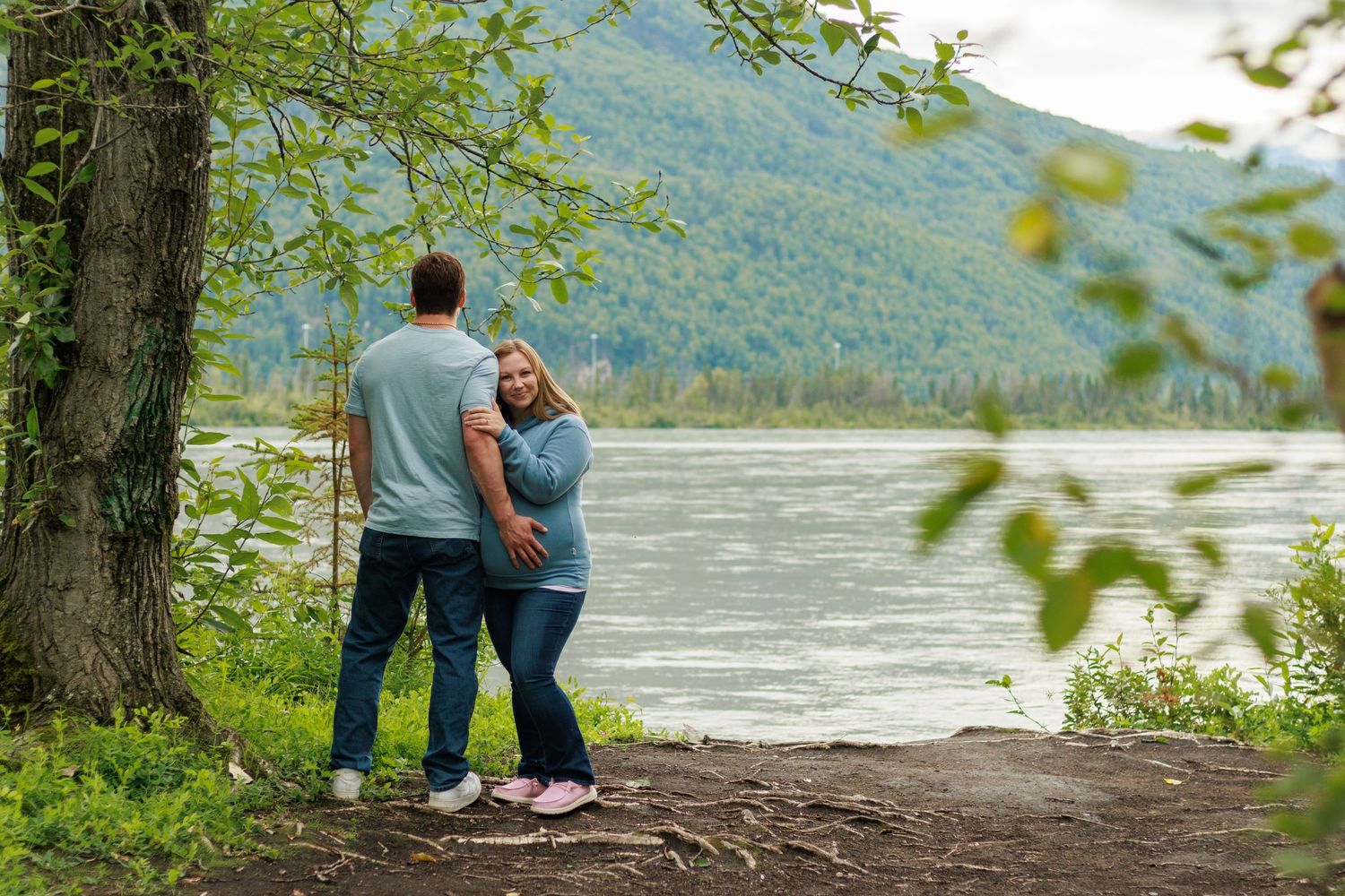 A couple stands together on a riverbank surrounded by green trees and mountains in the distance.