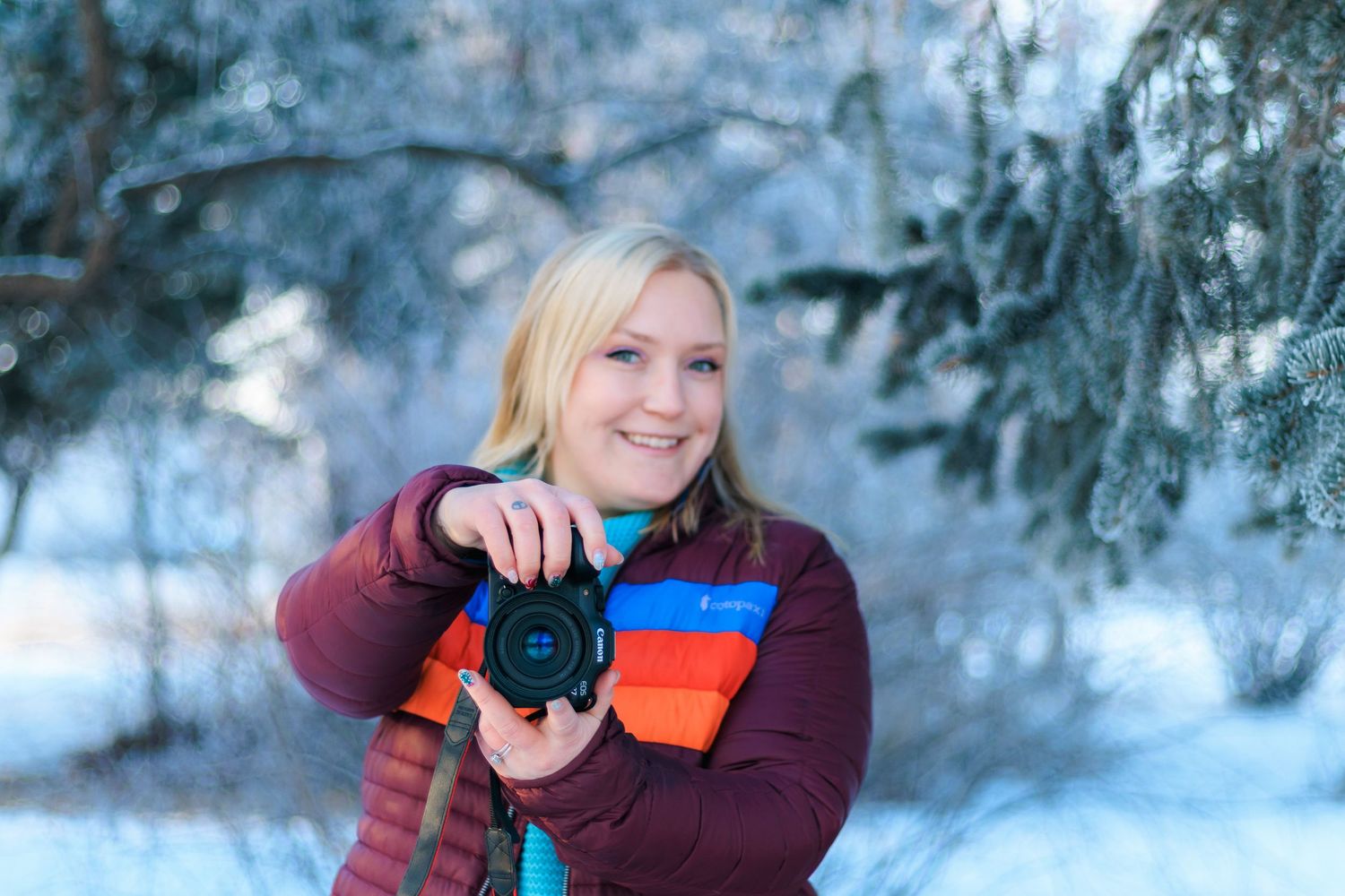 A photographer wearing a burgundy sweater takes pictures in a snowy winter forest setting with evergreen trees.