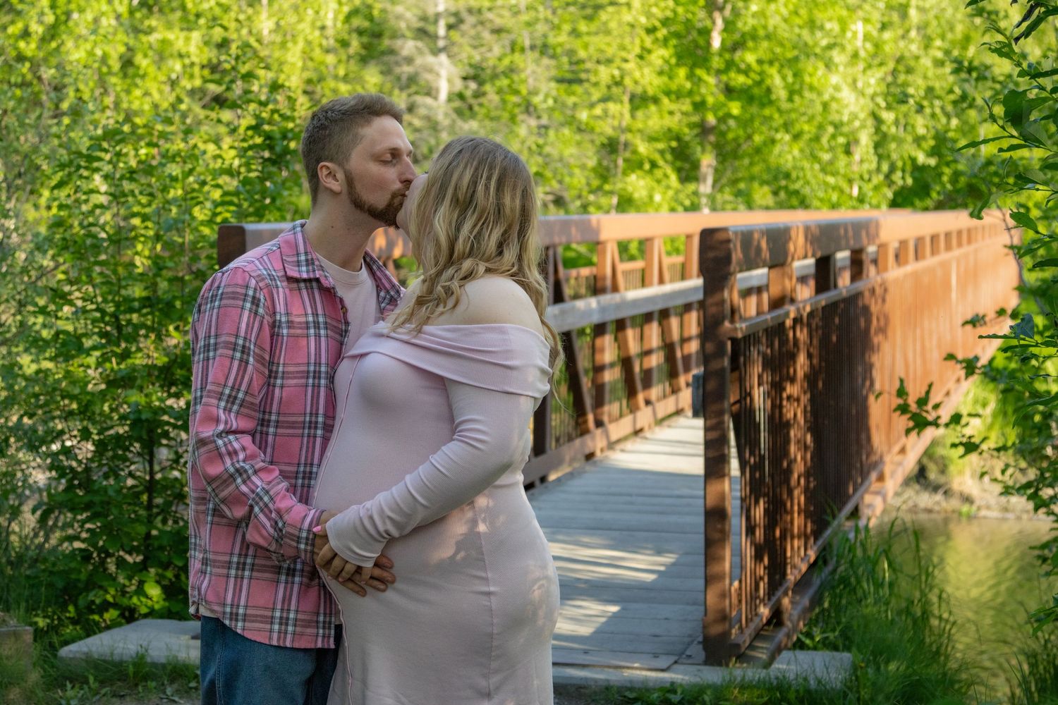 A couple shares tender moments together on a wooden bridge surrounded by lush greenery on a sunny day.