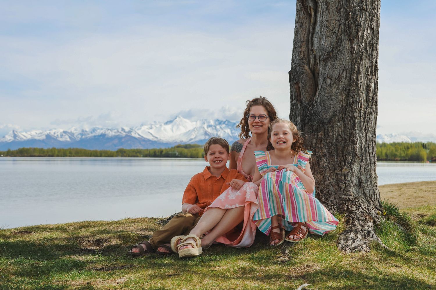 A group sits together under a tree by a lake with snow-capped mountains visible in the distance on a sunny day.