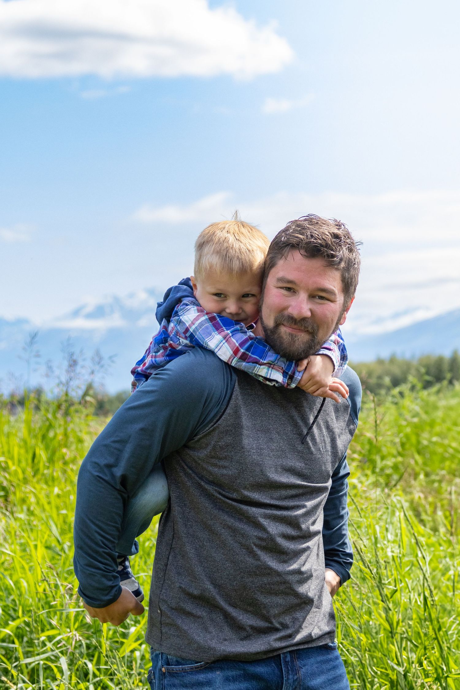 Person in gray shirt carrying child on shoulders while walking through sunny green meadow with blue sky.