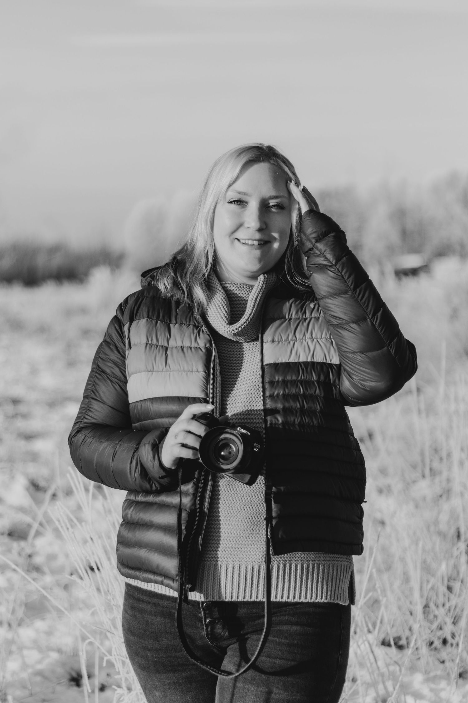 A photographer wearing a black puffer jacket holds a camera in a snowy winter field.