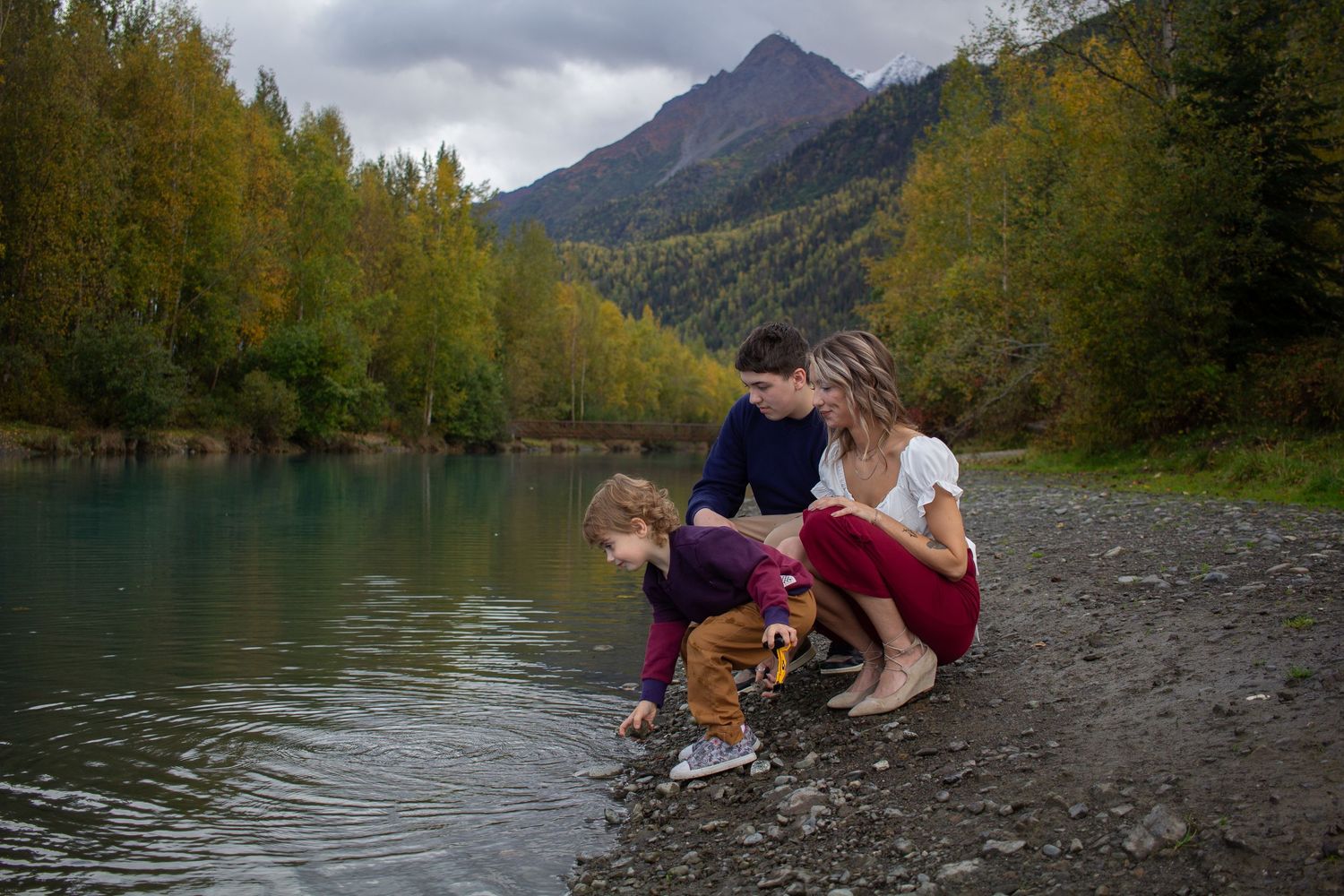 A scenic mountain lake surrounded by autumn trees with people skipping rocks along the rocky shoreline.