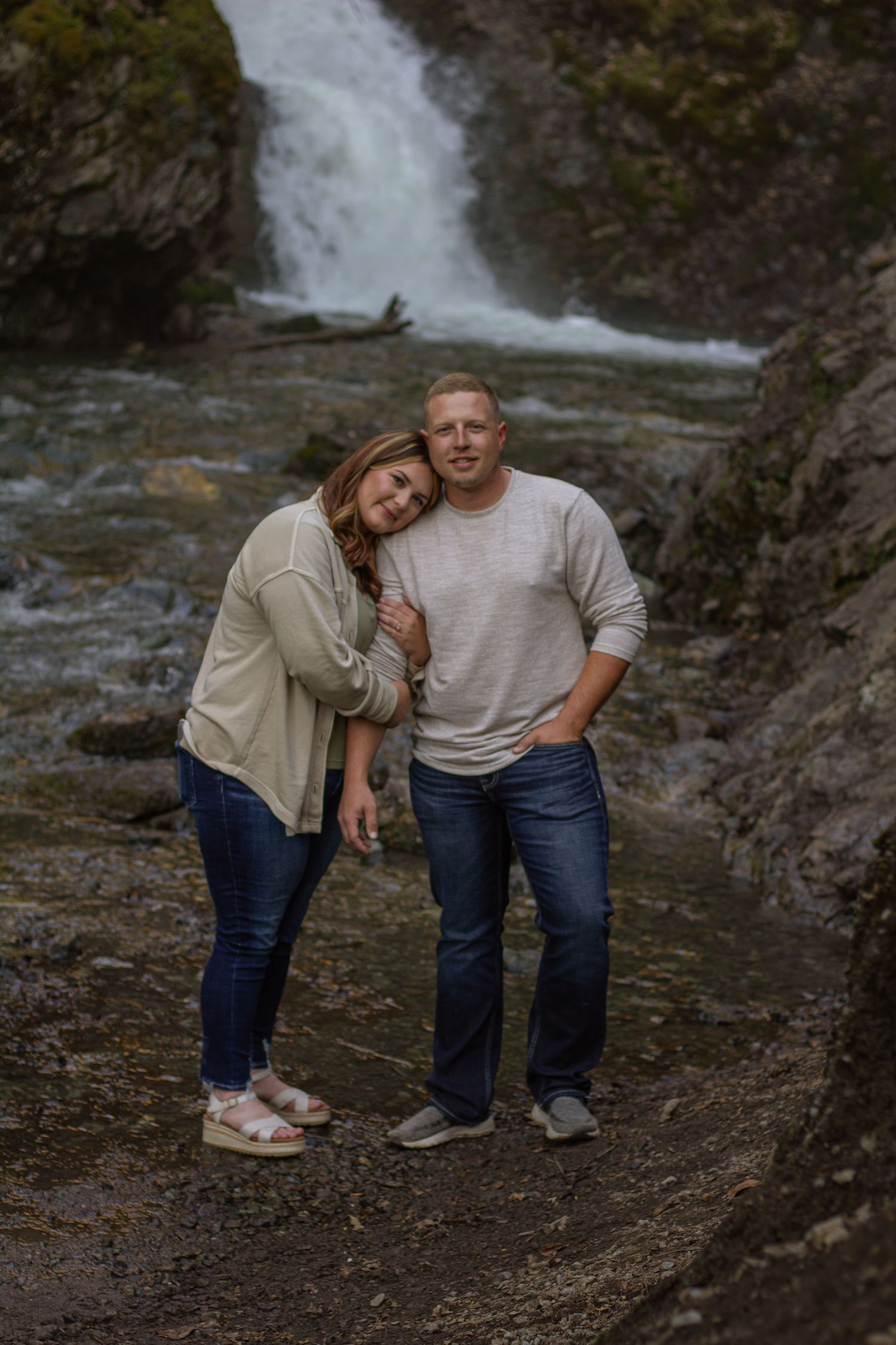 A couple shares an intimate moment together near a waterfall, dressed casually in jeans and light colored tops.