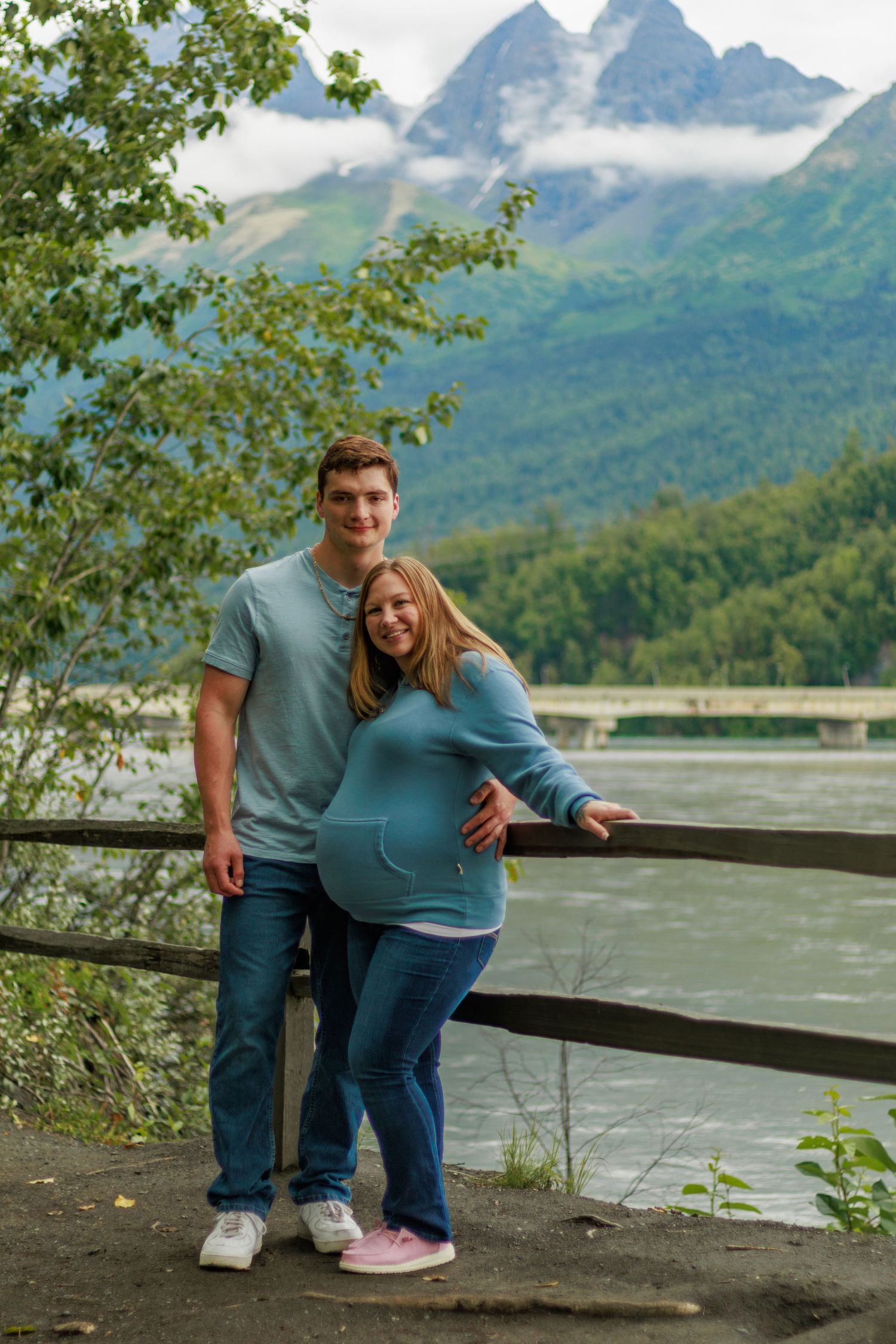A couple stands together on a scenic overlook with misty mountain peaks and a river in the background.
