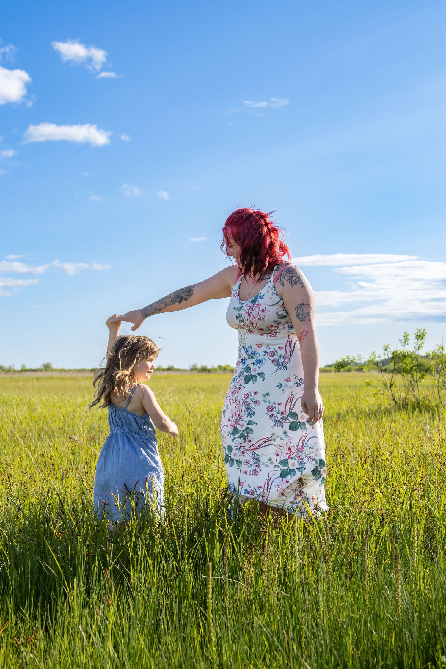 Two people in summer dresses walk through a lush green field under bright blue skies on a sunny day.