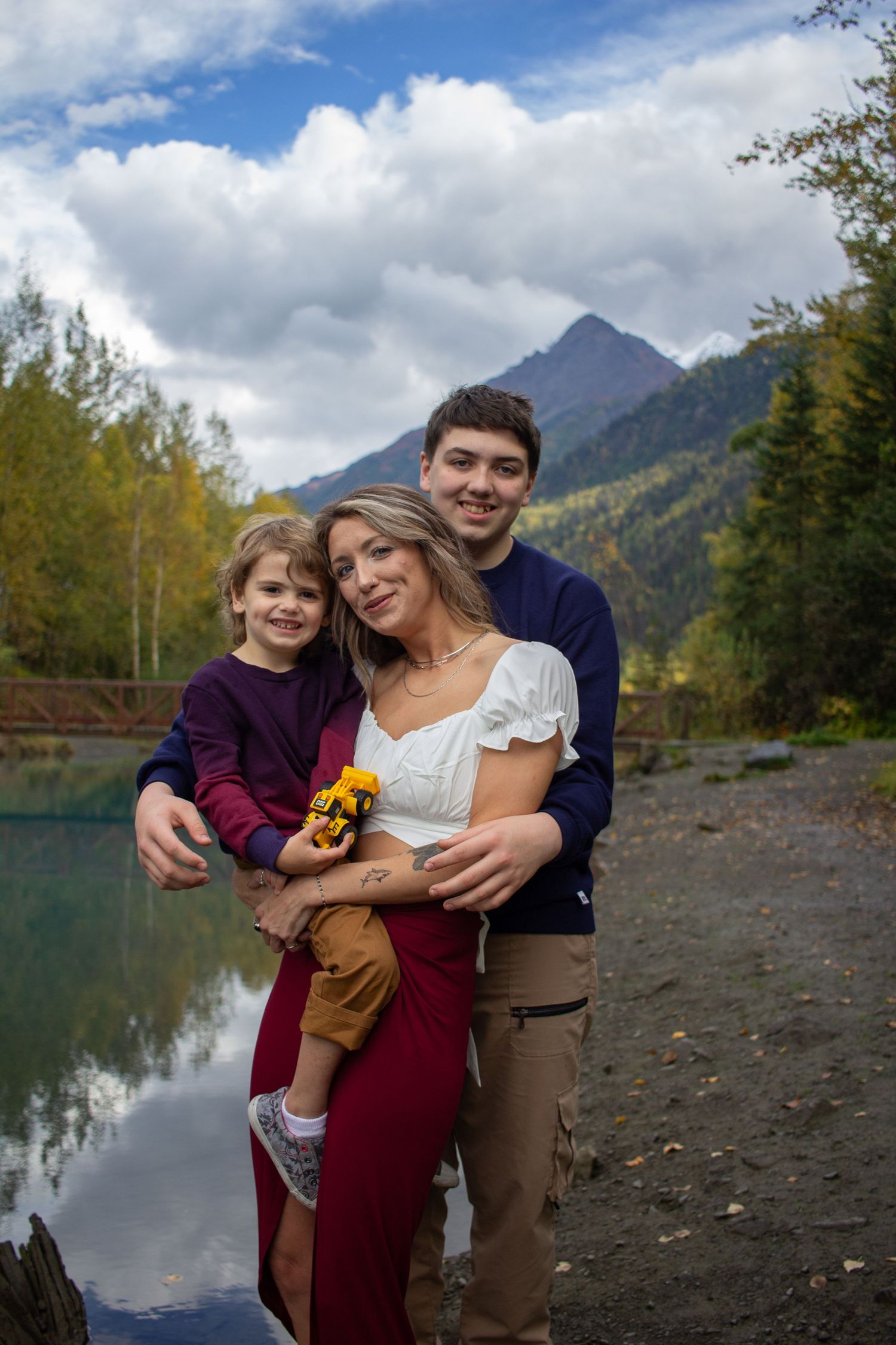 A family shares a tender moment by a serene mountain lake surrounded by autumn foliage and dramatic peaks.