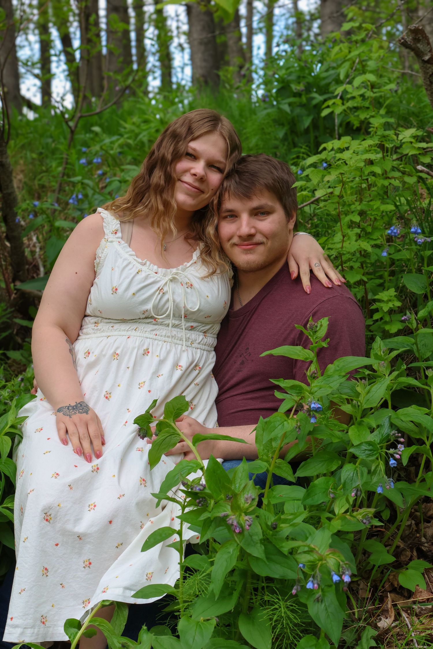 A couple poses together in a lush green forest setting wearing white and burgundy clothing.