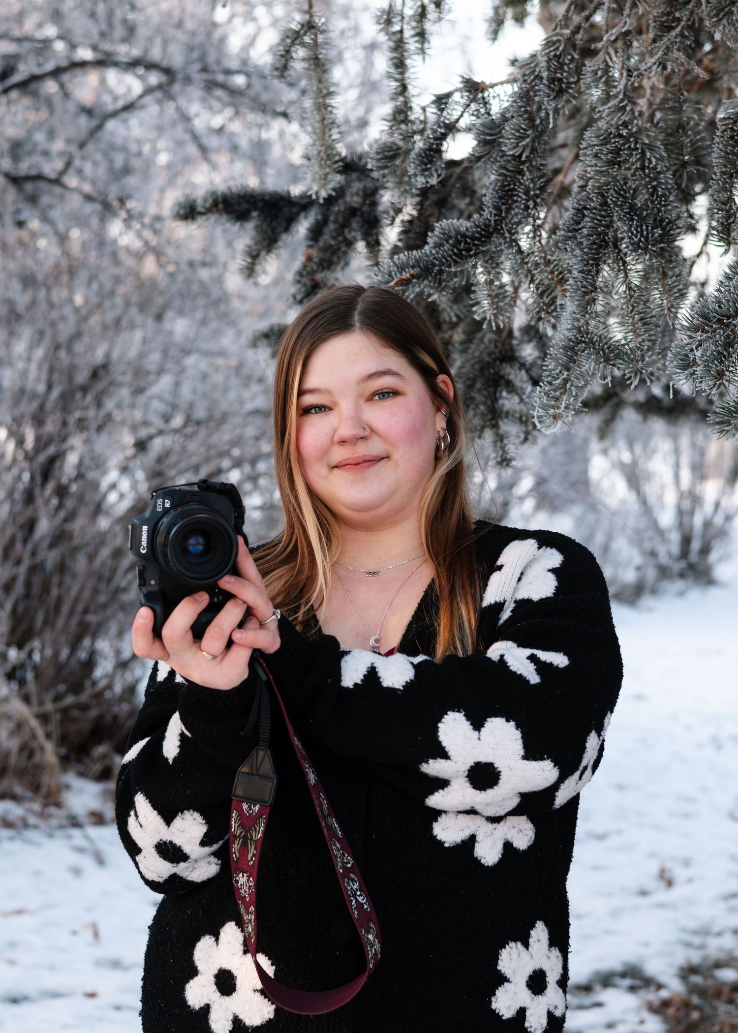 A photographer in a black and white floral sweater holds a camera in a snowy winter forest setting.