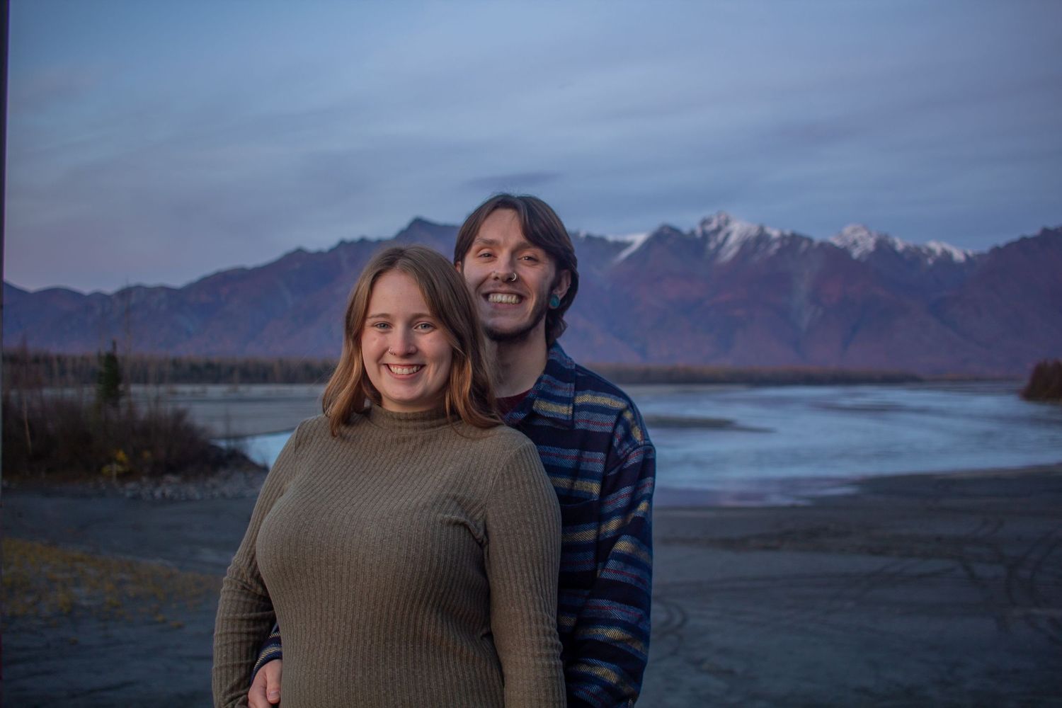 A couple stands together against a scenic mountain landscape at dusk with a lake in the background.