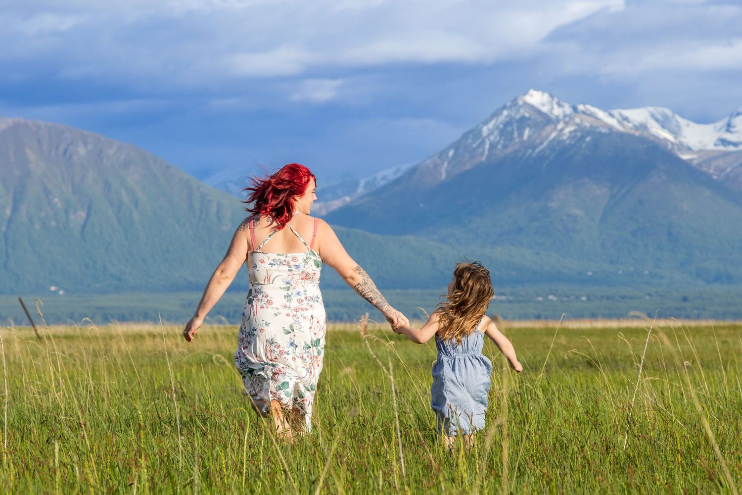 Two people walk through a scenic meadow with snow-capped mountains in the background on a sunny day.