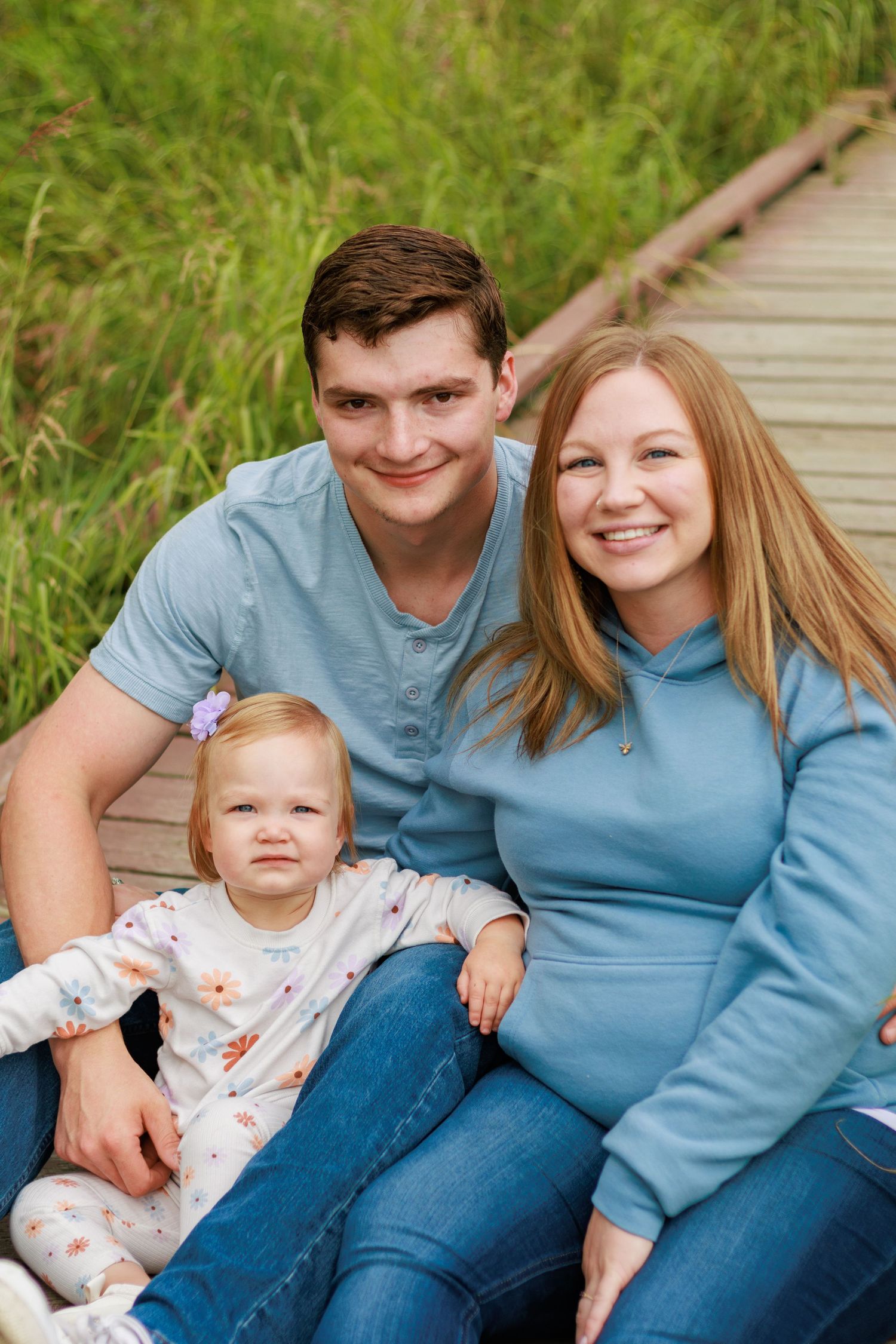 A family in coordinating blue outfits sits together on a wooden boardwalk surrounded by tall grass.