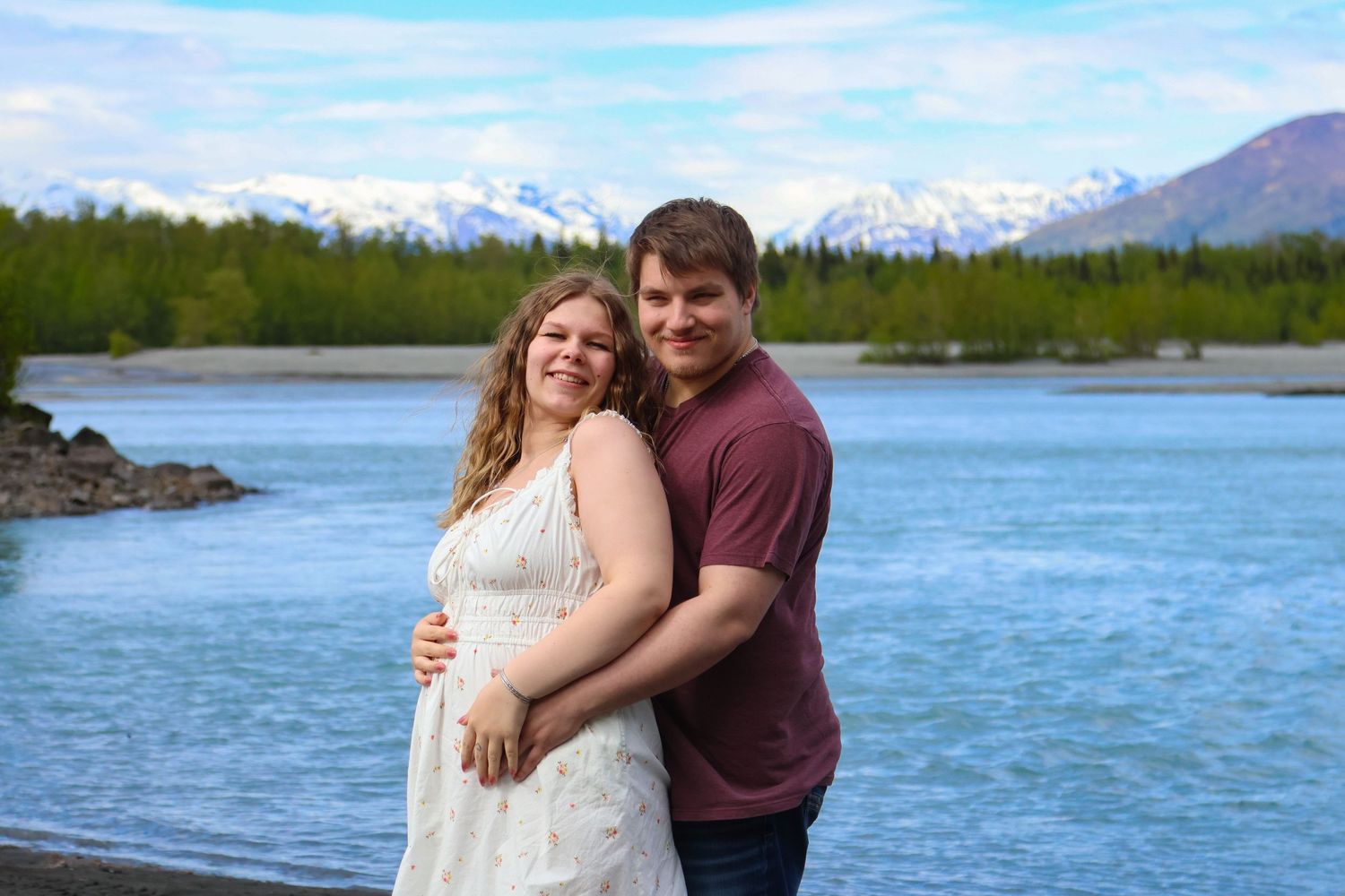Couple embraces against backdrop of snow-capped mountains and turquoise lake in scenic Alaskan landscape.