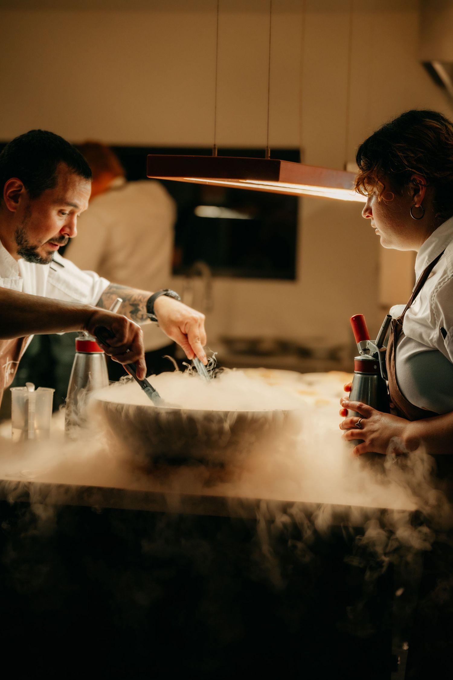 Two chefs use liquid nitrogen in a restaurant kitchen, creating dramatic smoke effects over a large bowl.