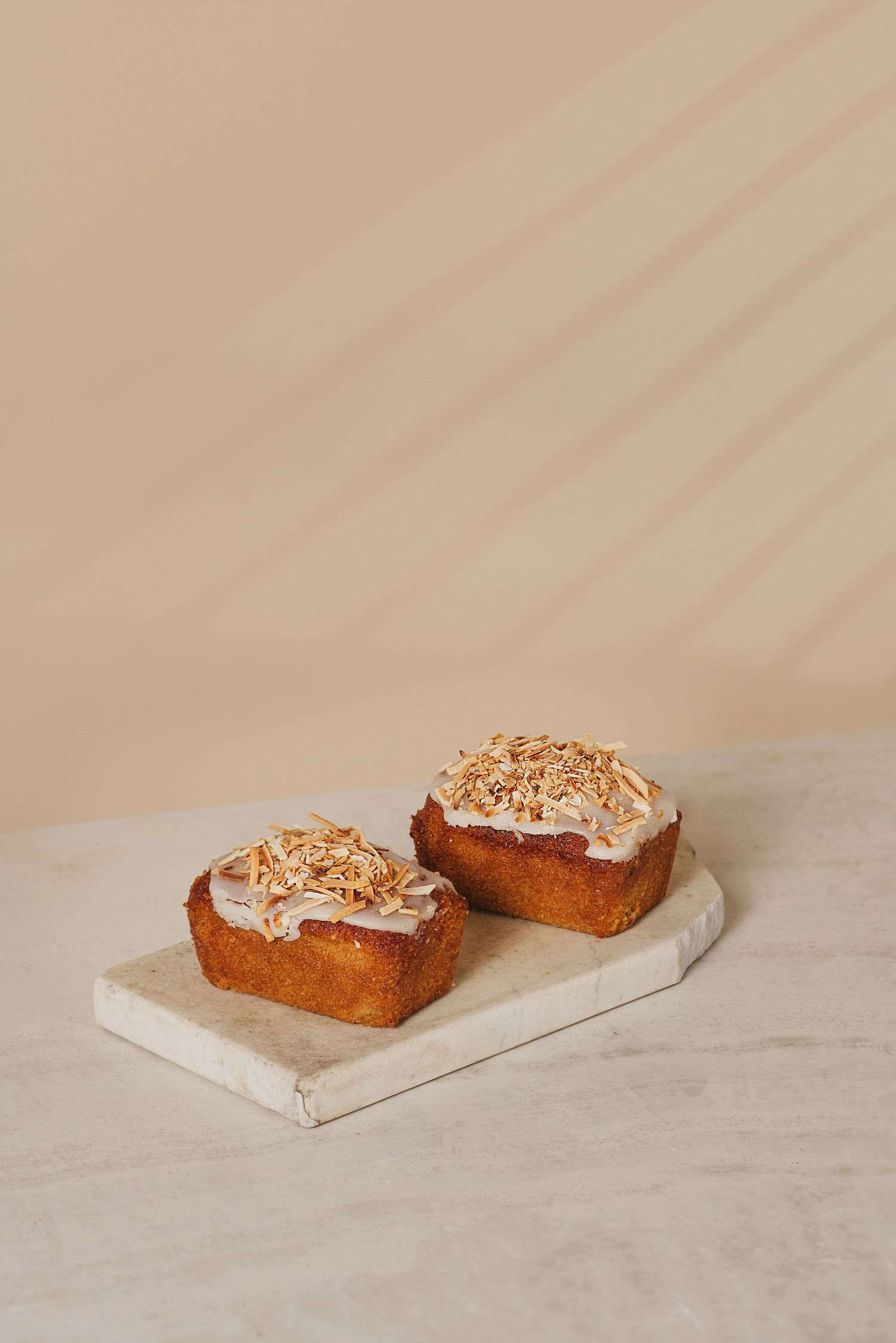 Two frosted mini loaf cakes topped with toasted coconut flakes on a marble serving board.