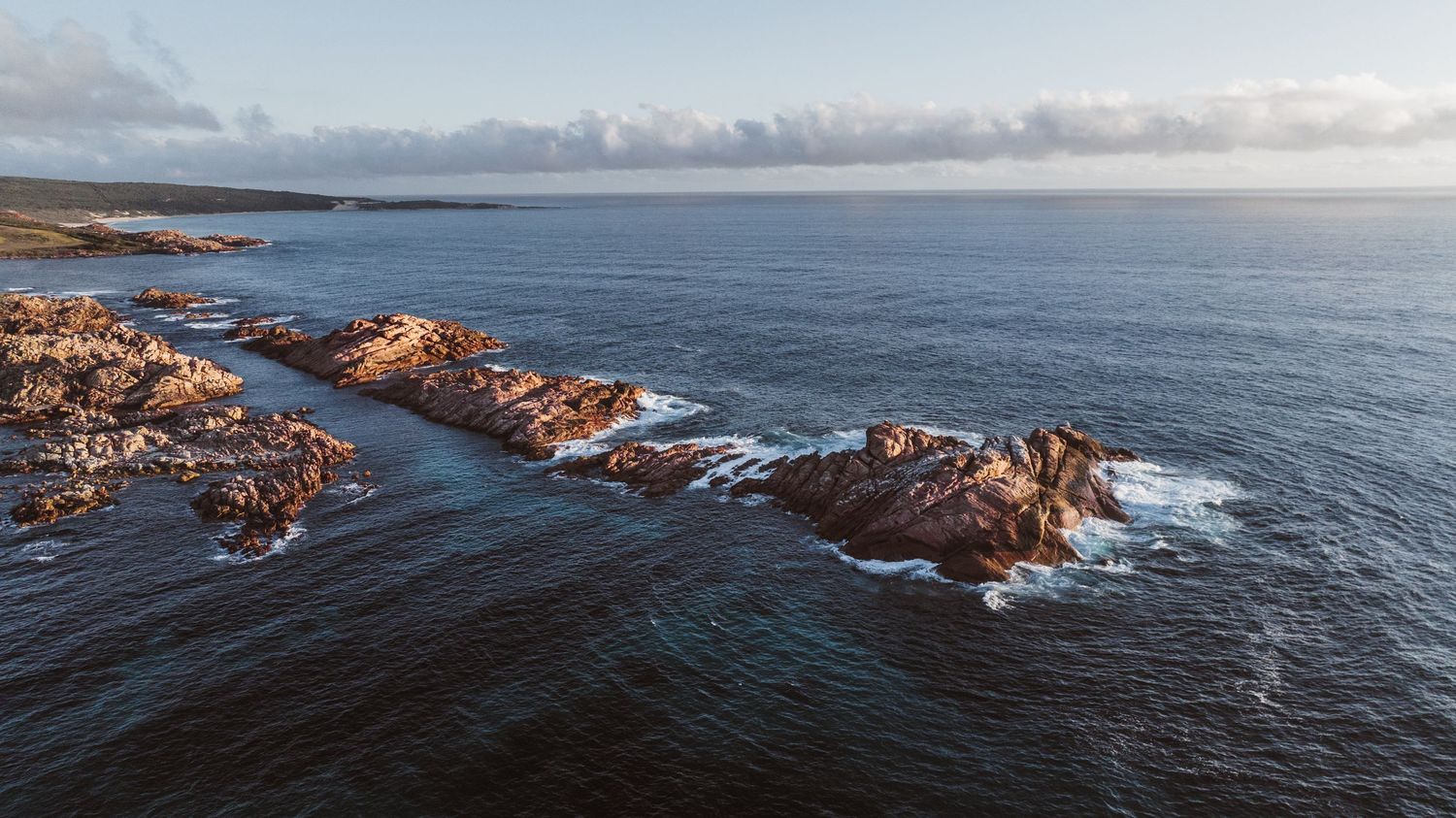 Aerial drone view of rocky coastal outcrop surrounded by calm blue ocean waters at dusk.