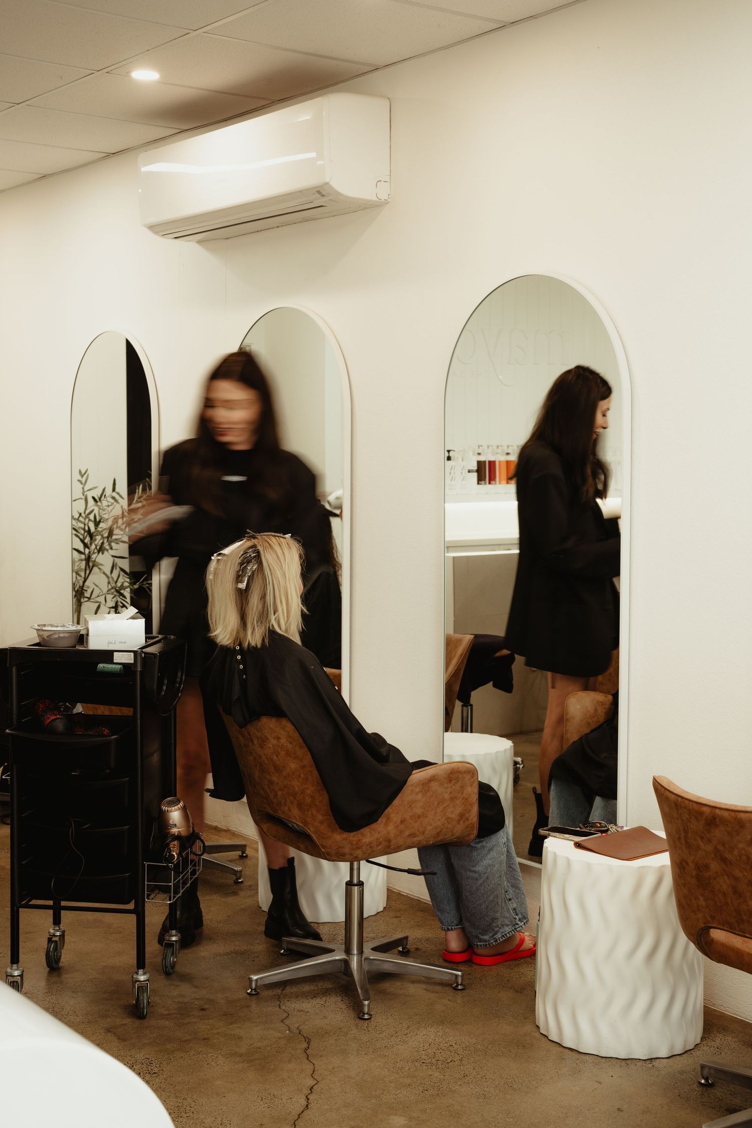 Hair stylist blowdrying blonde clients hair in a bright modern salon with arched mirrors.