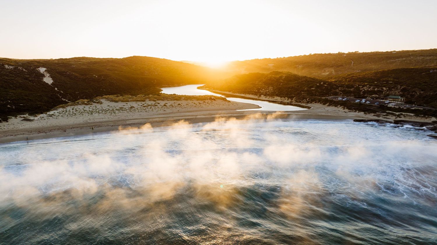 Aerial view of golden sunset over coastal beach with waves and rolling hills in background.