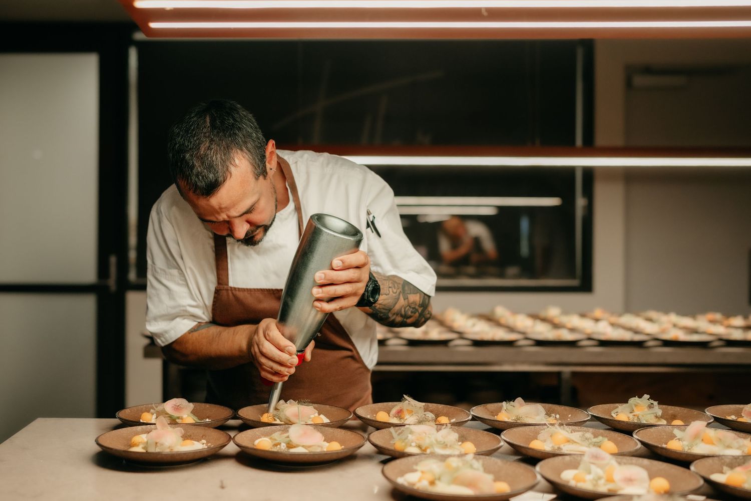 Chef carefully piping cream onto multiple plated desserts in a professional restaurant kitchen.