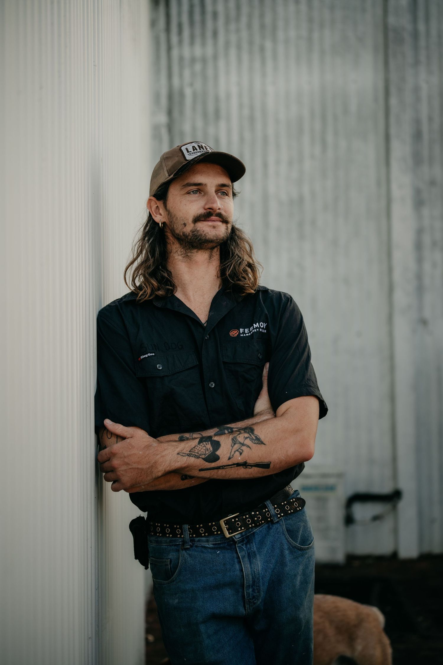 Tattooed man with long hair and cap leaning against a corrugated wall with arms crossed outdoors.