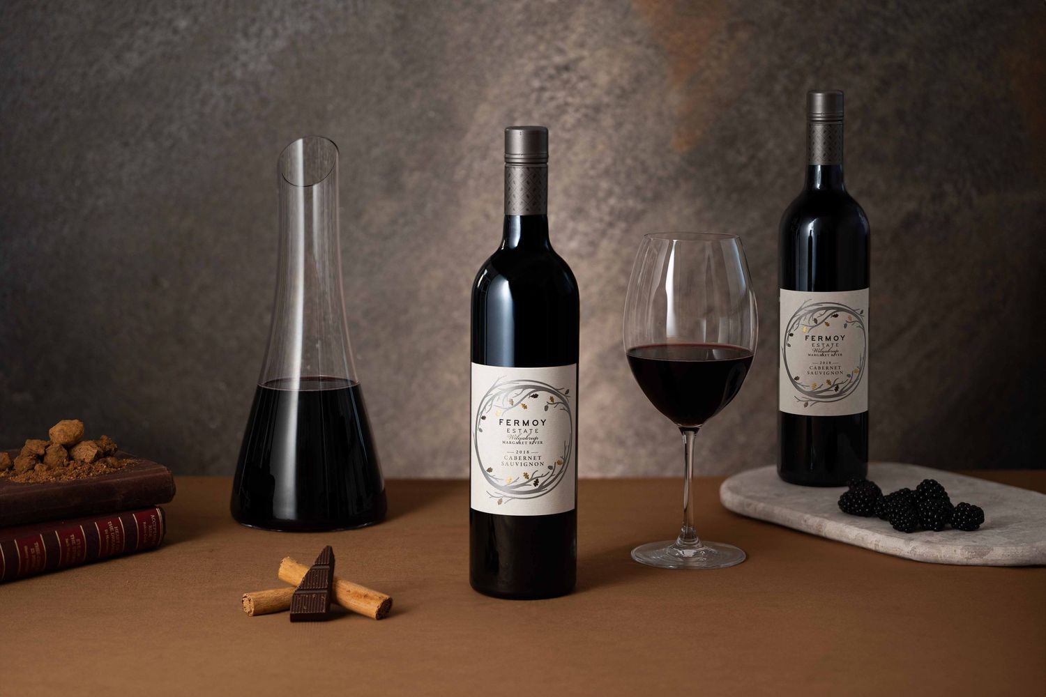 Fermoy Estate red wine bottles displayed with a glass, decanter, chocolate and cork on a table.