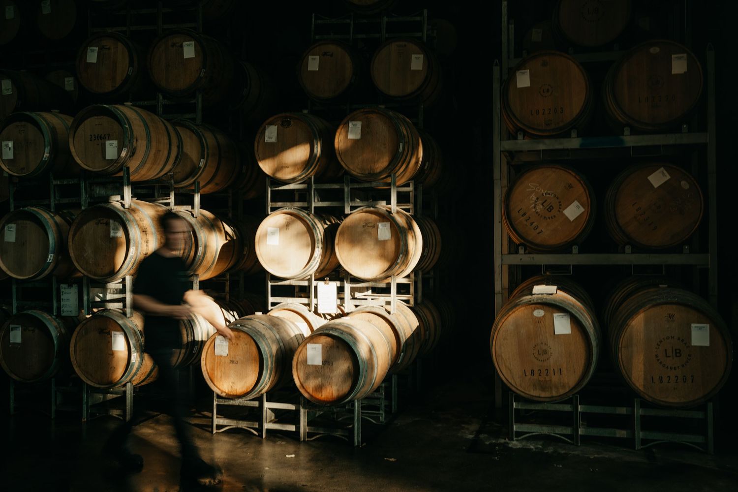 Worker inspecting oak wine barrels stacked on metal racks in a dimly lit winery cellar.