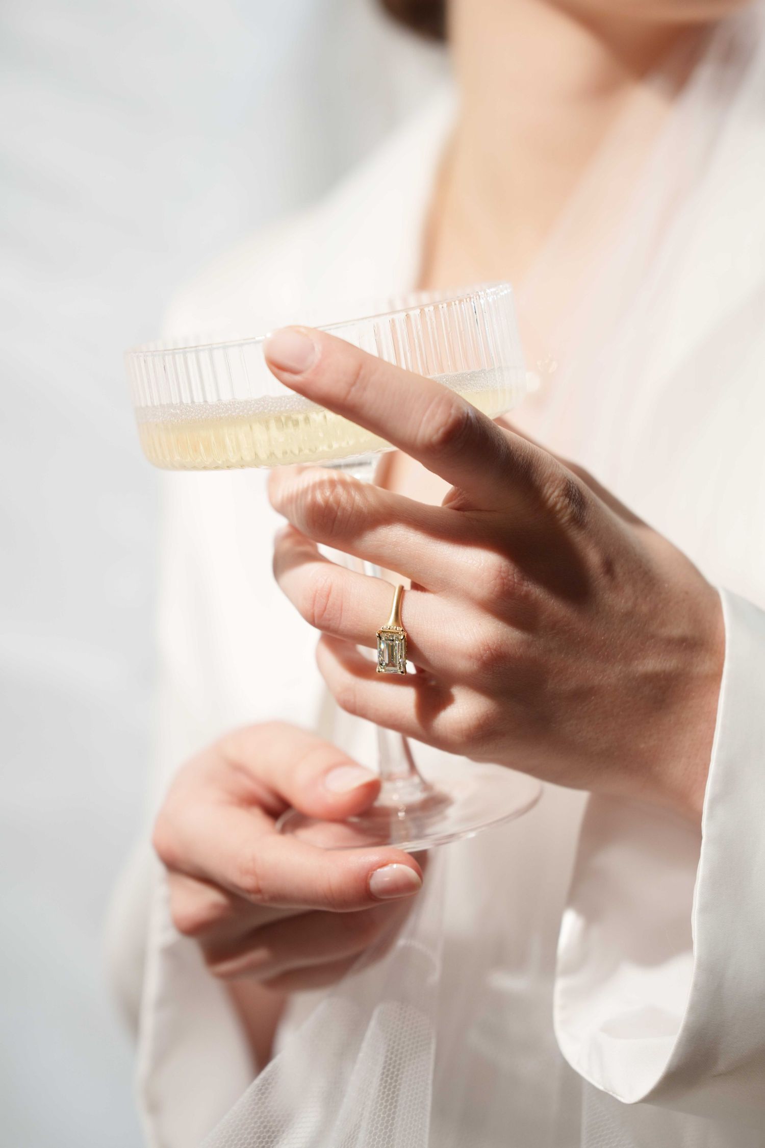 Close-up of a woman in a white robe holding a ribbed coupe glass of champagne with a diamond ring.