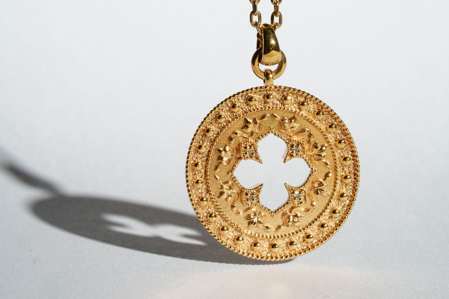 Gold circular pendant necklace with an ornate quatrefoil cutout design casting a shadow.