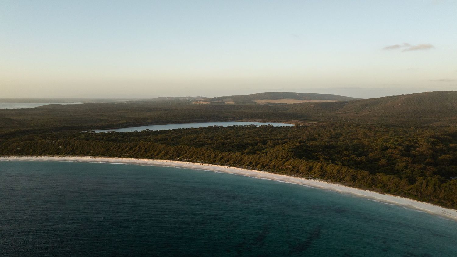Aerial coastal landscape featuring a white sandy beach, turquoise water, and a serene lake nestled in forest.
