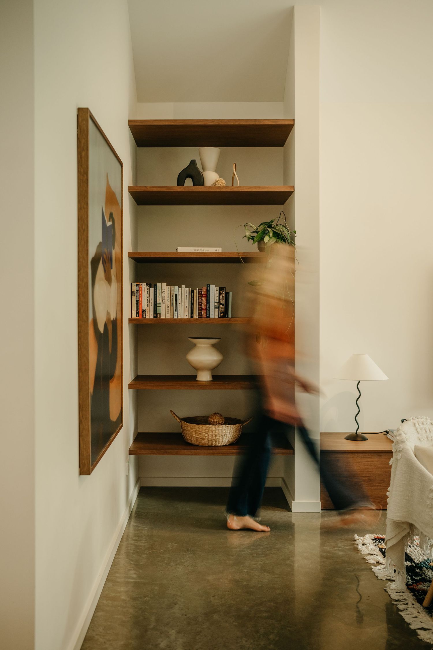 Blurred figure walks past built-in wooden shelves with books and decor beside abstract wall art in modern home.