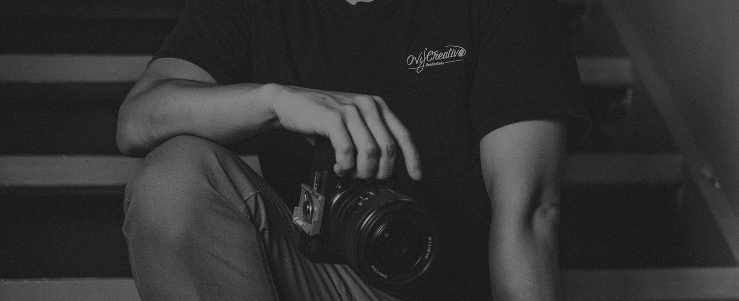 Black and white photo of a photographer sitting on steps holding a DSLR camera in casual attire.