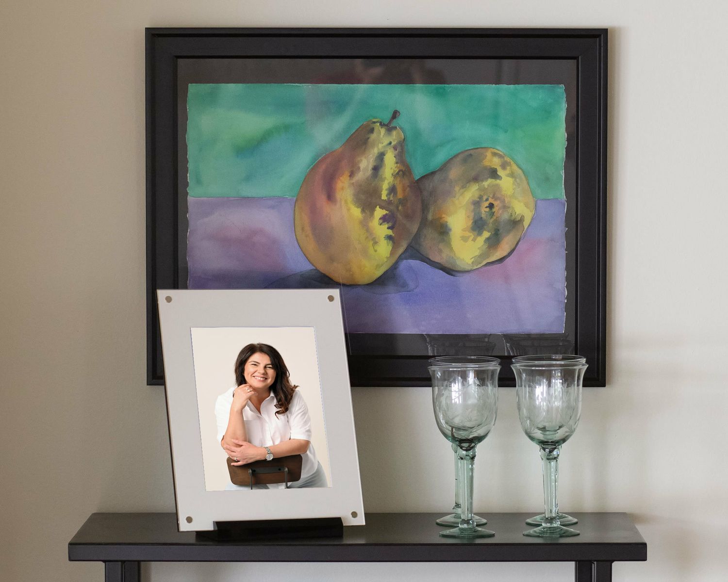 White framed woman portrait on black console table beside wine glasses below a colorful pear painting.