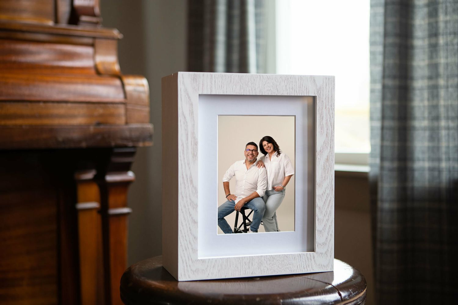 White textured picture frame with couple portrait displayed on a round wooden side table near a window.