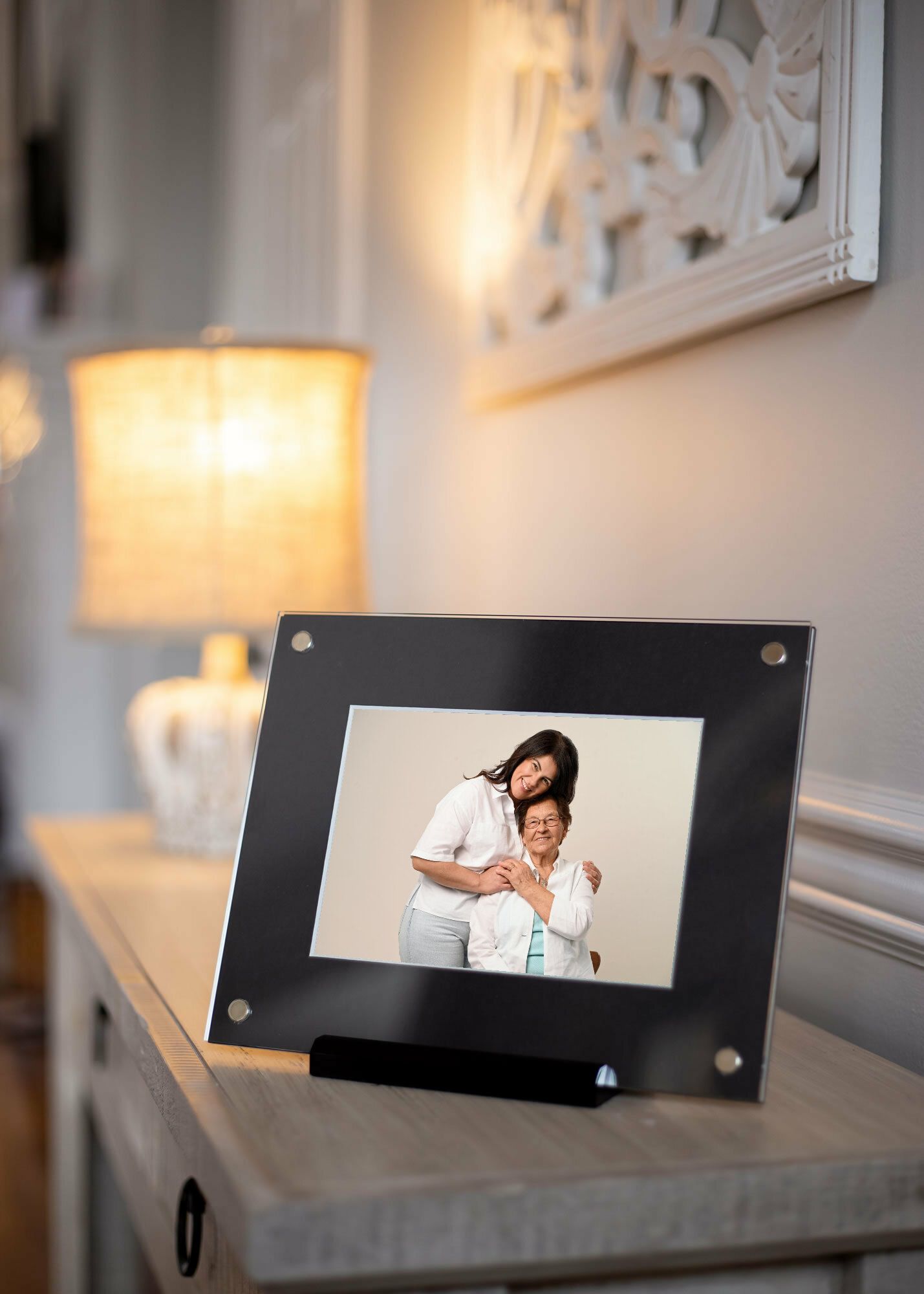Black acrylic framed portrait of two women on a gray console table beside a glowing lamp and ornate wall art.
