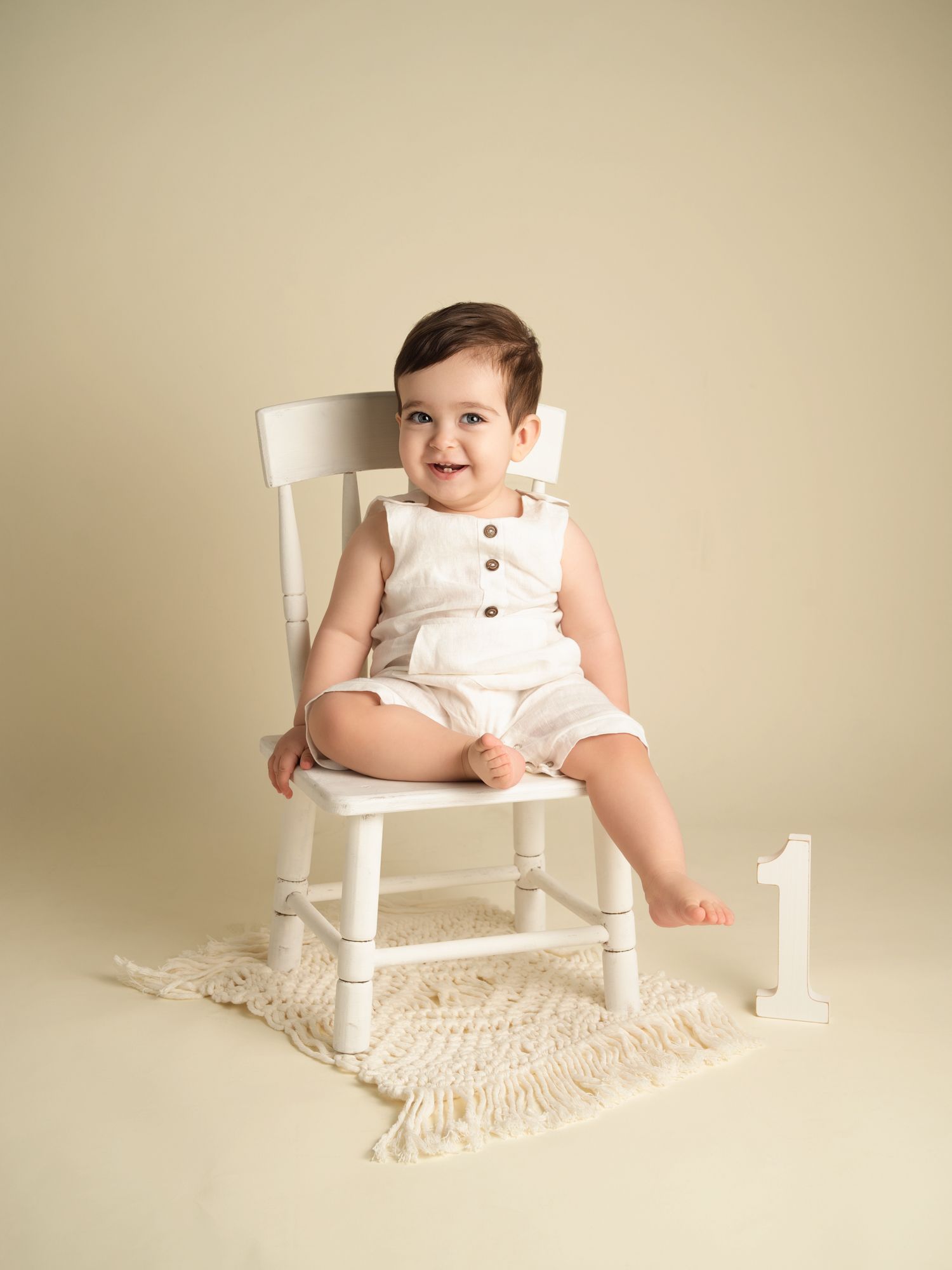 Smiling baby in white romper sitting on a white chair next to a number one, milestone photo.