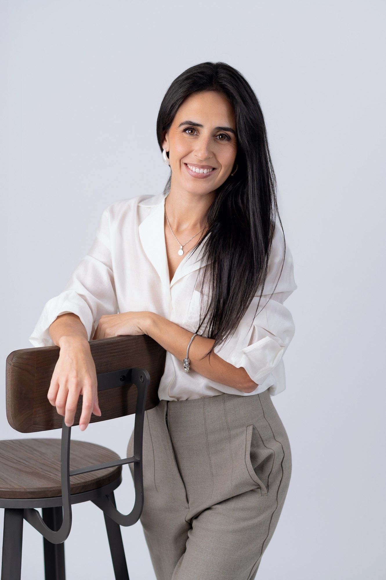 Smiling woman with dark hair in white blouse and gray trousers leaning on a wooden bar stool against light background.