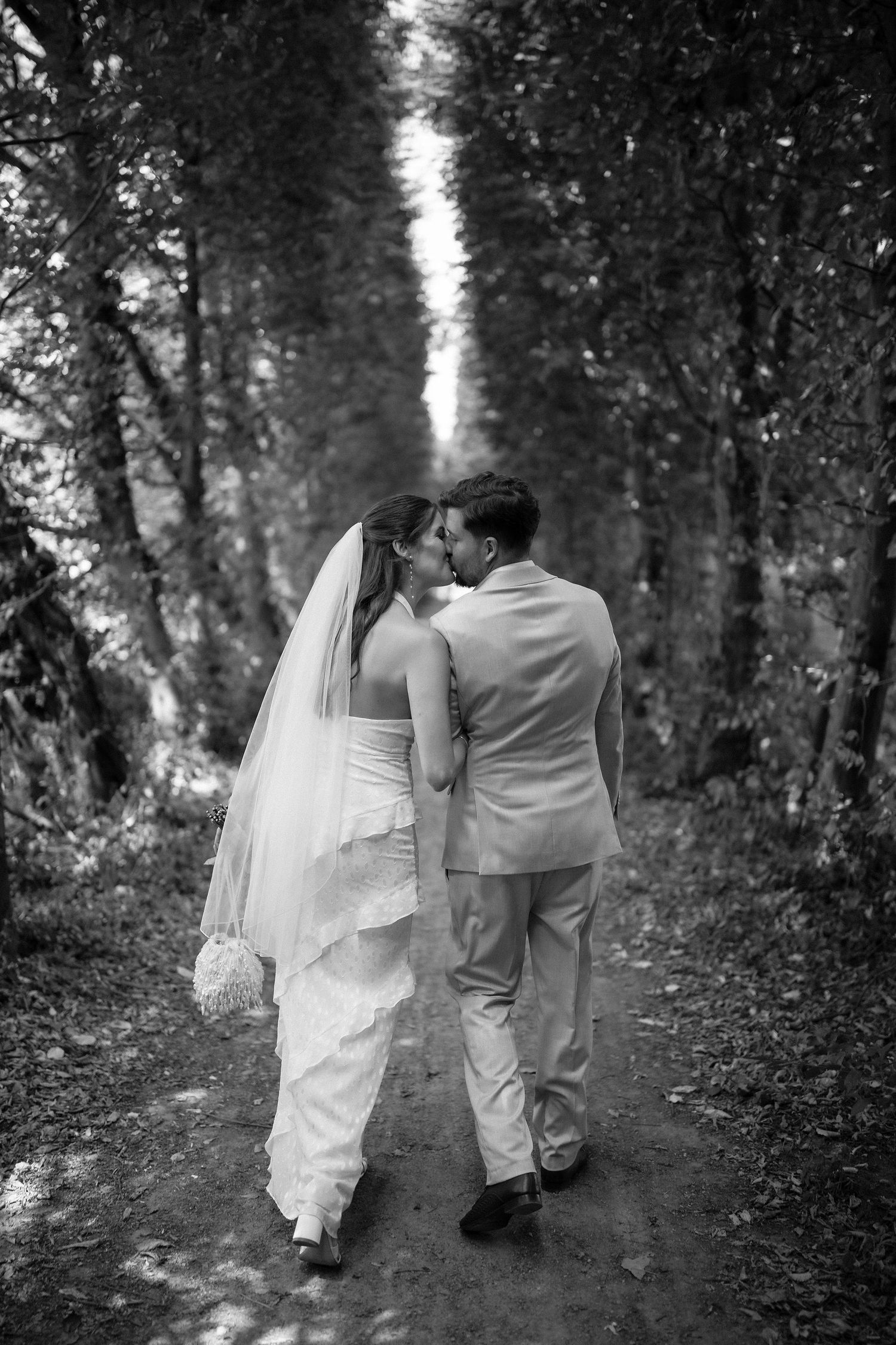 Black and white photo of a bride and groom kissing while walking down a tree-lined path on their wedding day.
