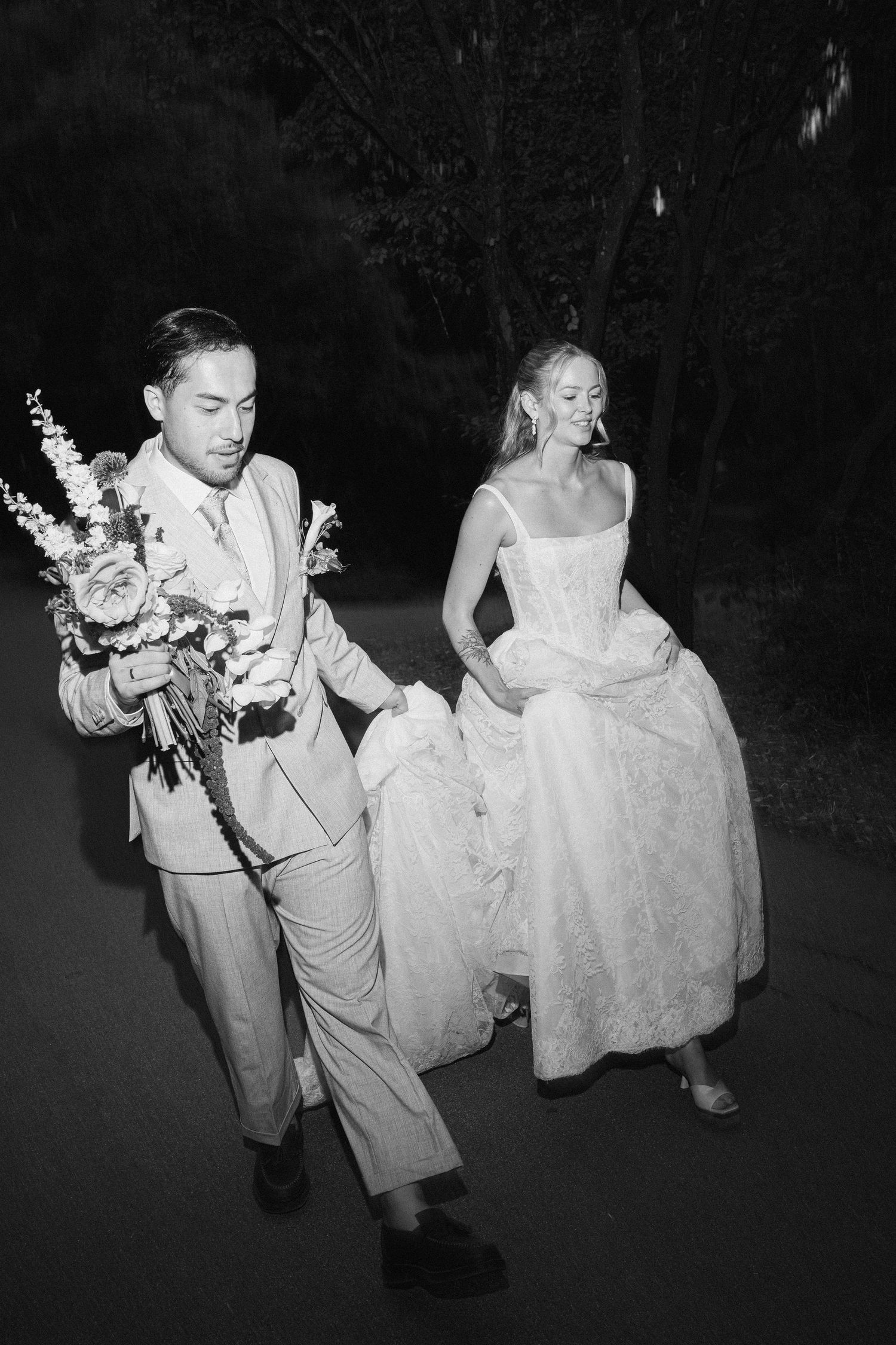 Black and white photo of bride and groom walking outdoors at night, groom holding bridal bouquet.