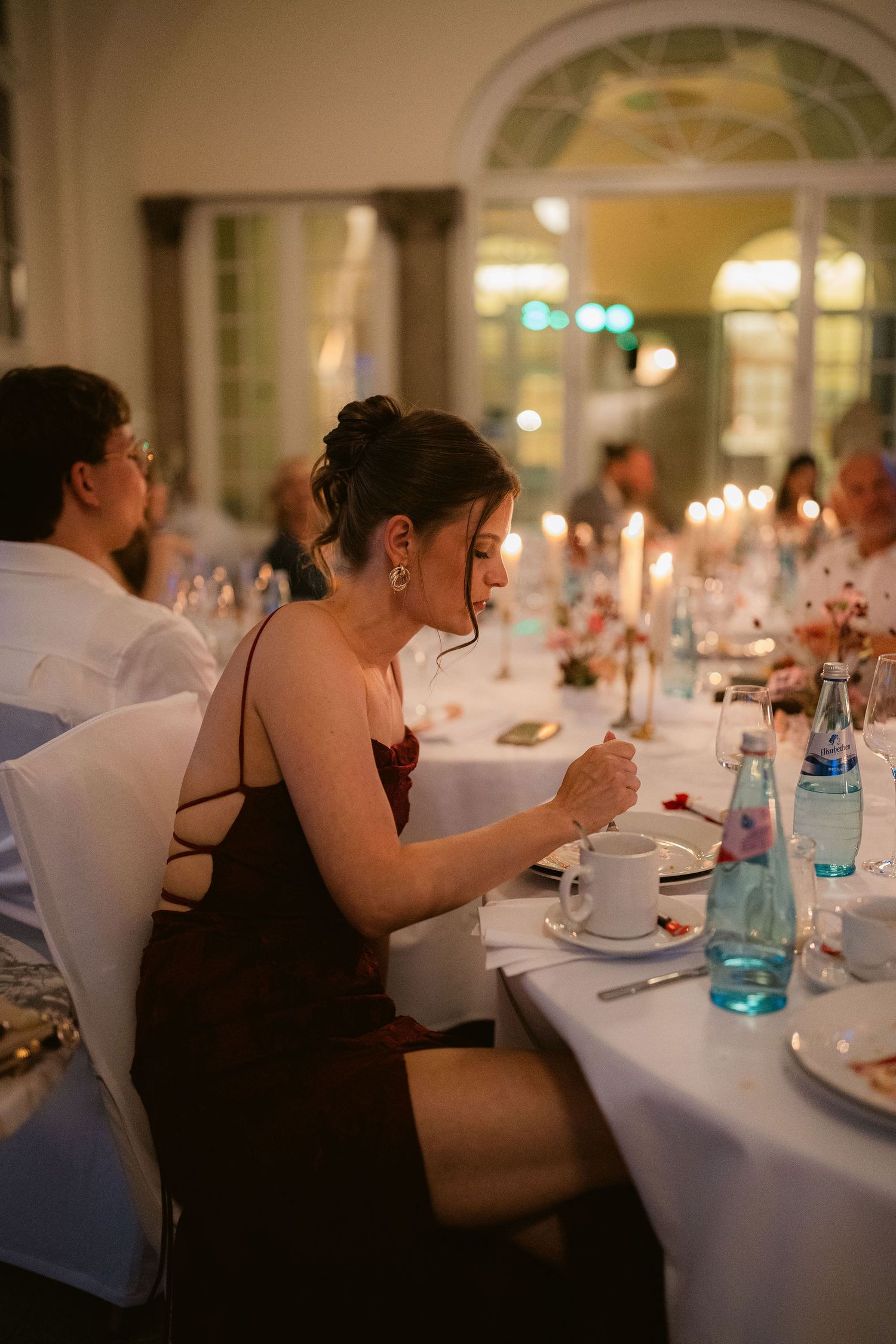 Woman in burgundy dress seated at candlelit dinner table during elegant evening reception.