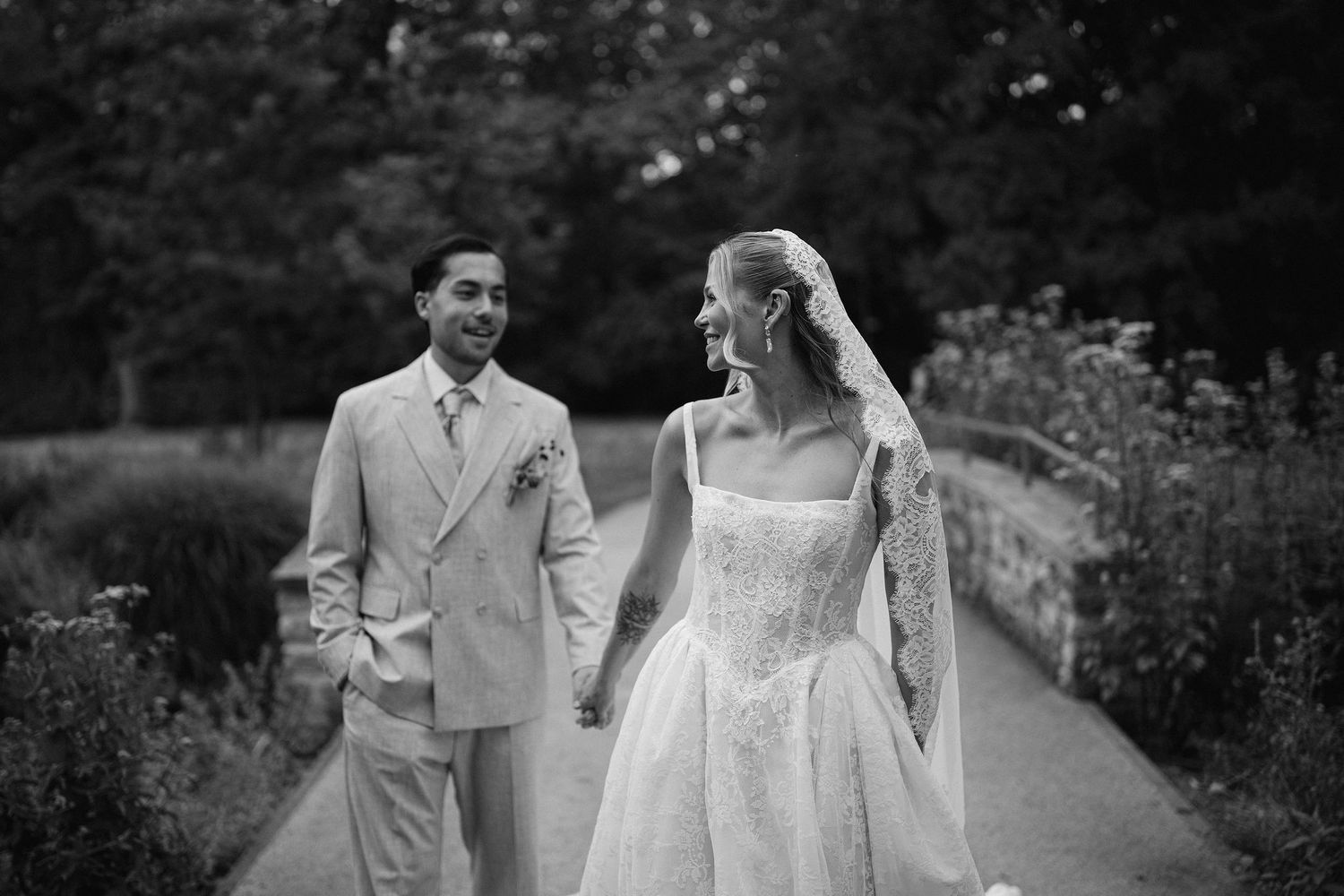 Black and white photo of a bride and groom holding hands, walking on a path surrounded by lush greenery.