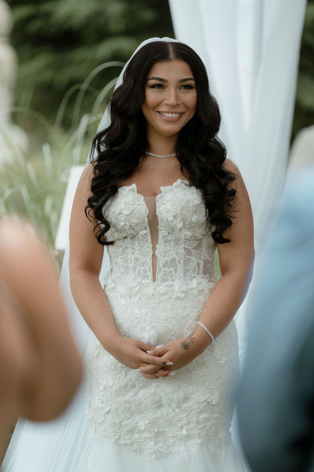 A bride in a stunning white lace wedding gown poses for portraits with dark flowing hair and elegant jewelry.