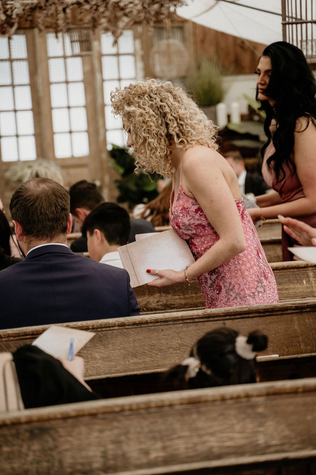 Wedding guests sit in wooden church pews during ceremony, with soft brown tones and natural lighting.