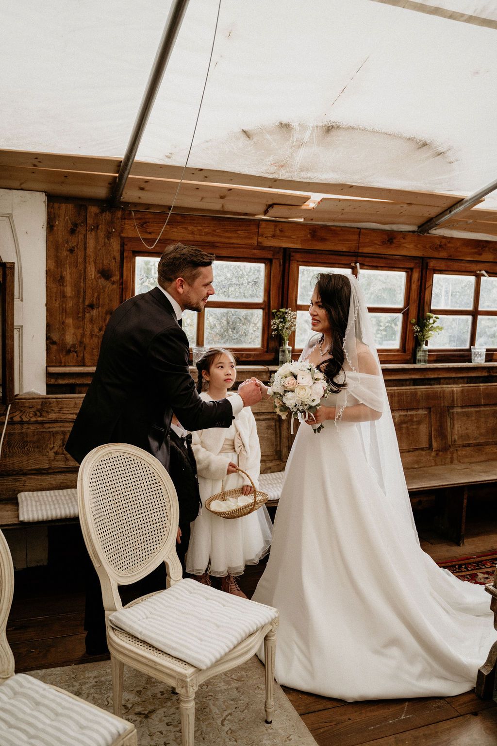 A romantic rustic wedding reception scene showing a couple seated at a wooden table with white linens and floral decor.