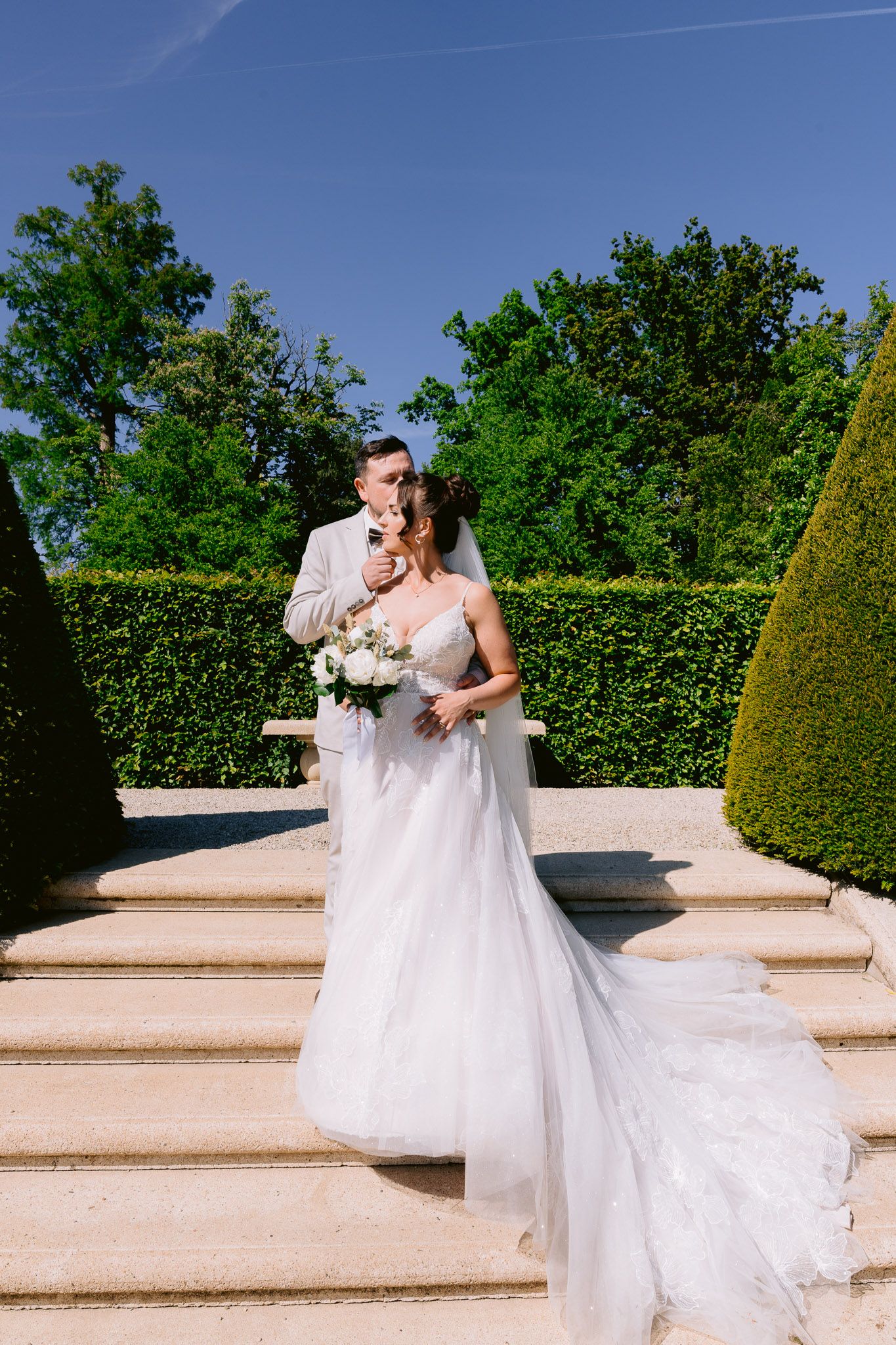 A couple shares a tender moment on white stone steps in a manicured garden with lush hedges and topiary under blue sky.