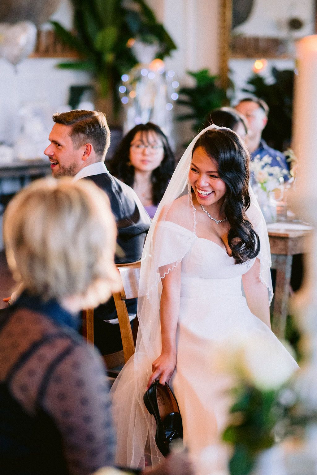 A joyful bride wearing a white off-shoulder wedding dress and veil laughs during an intimate indoor wedding reception.