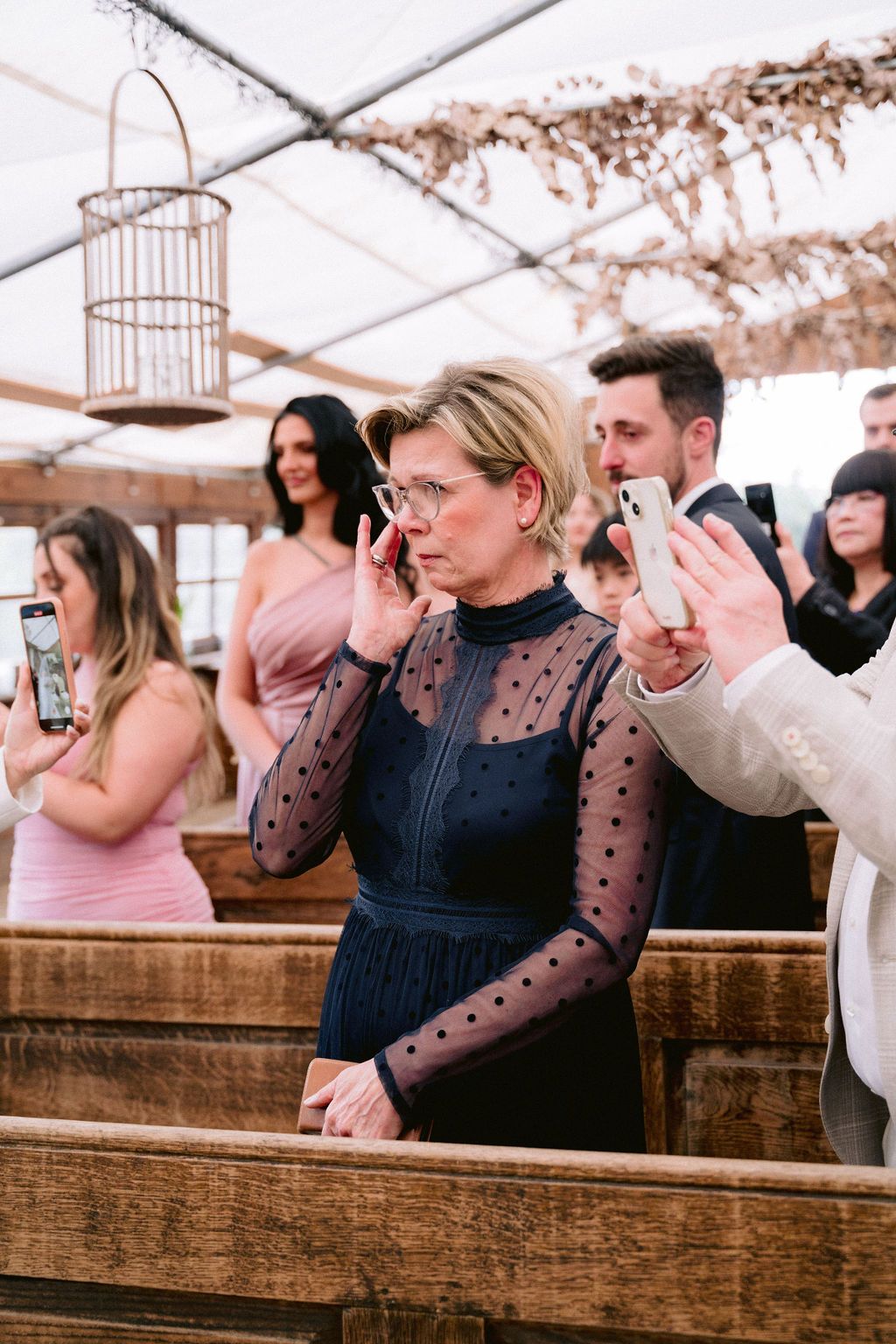 Guests stand at rustic wooden benches under a tent during an event, with a decorative birdcage hanging above.