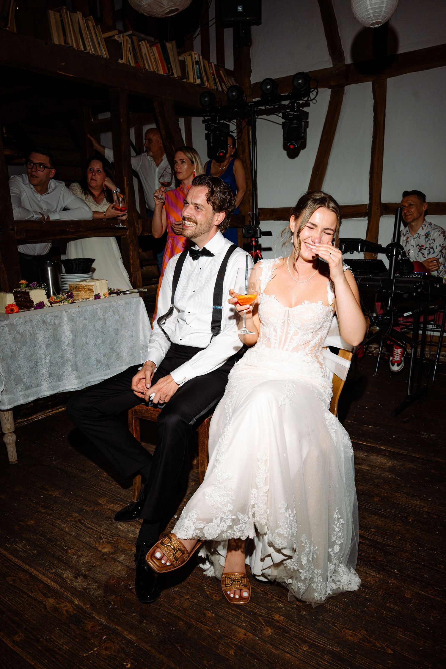 Bride and groom laughing together at wedding reception, seated in rustic venue with guests in background.