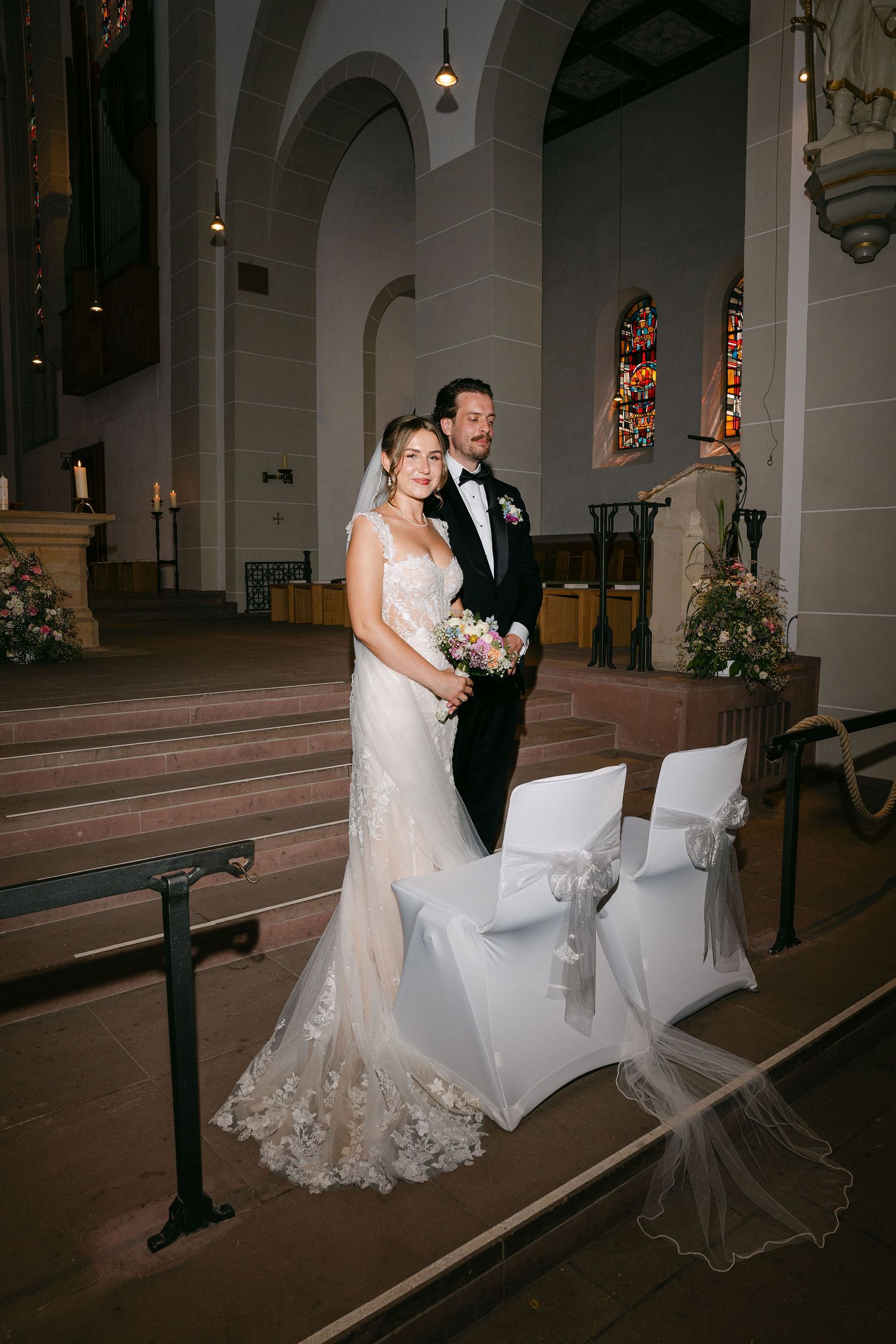 Bride and groom posing together at church altar, bride in lace mermaid gown with veil, groom in black tuxedo.