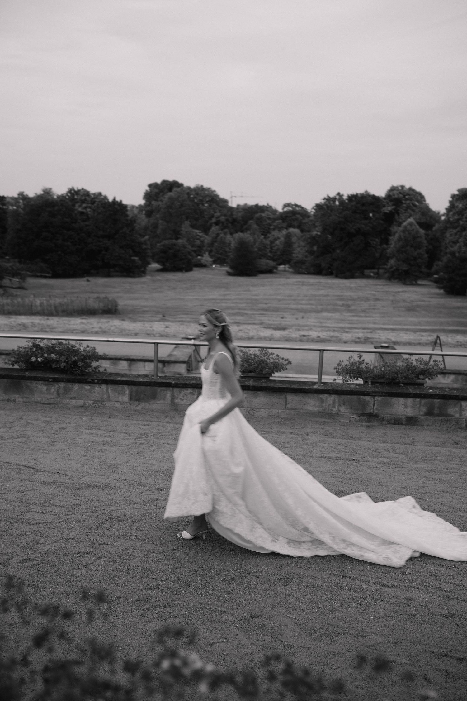 Black and white photo of a bride in a flowing wedding gown with long train walking on an outdoor terrace.